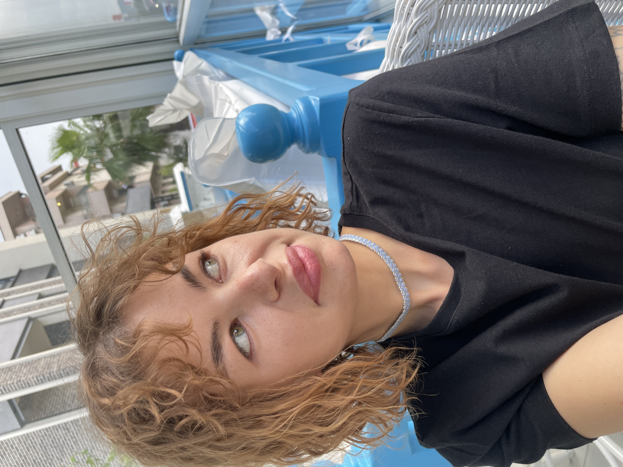 A medium, angled close-up shot shows a woman with fair skin, green eyes, and curly light brown/blonde hair wearing a black t-shirt and a light blue and white necklace. The shot is taken in a location with a blue and white architectural structure, possibly a gazebo or enclosed patio. Outside the structure, in the background, is a partial view of a tall building and a palm tree. This could possibly be Podgorica, Montenegro. The woman is looking slightly upward and to the left of the camera, with a neutral expression. The background is somewhat blurred.