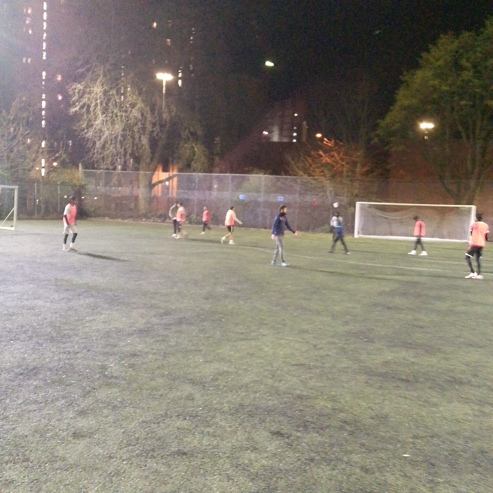 An overhead, low-light photograph depicts an active nighttime football match or practice on an artificial turf pitch in Coventry, United Kingdom. Approximately ten individuals, primarily males, are distributed across the field, wearing various athletic attire including orange, white, blue, and dark colored tops. Two white football goals with nets are present at opposing ends of the pitch. A football is visible airborne near the right side of the frame. The field is illuminated by several overhead floodlights, casting shadows. In the background, beyond a chain-link fence, bare trees and illuminated windows of multi-story buildings are discernible, indicating an urban setting.