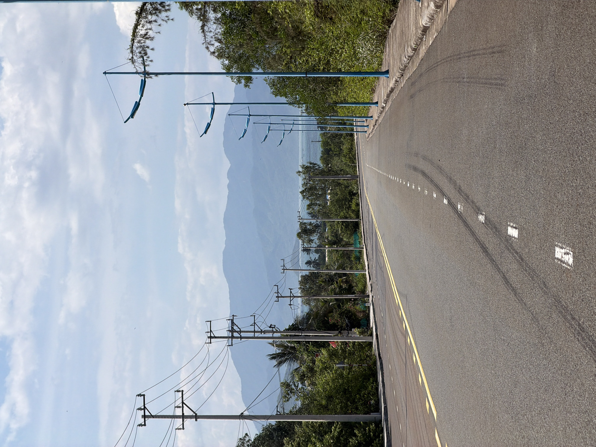 This is a vertical, eye-level, medium shot of a paved road in Vĩnh Lương, Vietnam, with a mountainous landscape in the background. The road has a yellow double line marking its center and white dashed lines marking the right edge. Tire marks are visible on the asphalt. On the left side of the road, there are multiple utility poles with wires. On the right side, there is a sidewalk with a row of distinctive blue lampposts. Lush green trees and vegetation line the right side of the road. The sky is partly cloudy. There are no people or animals visible in the image.