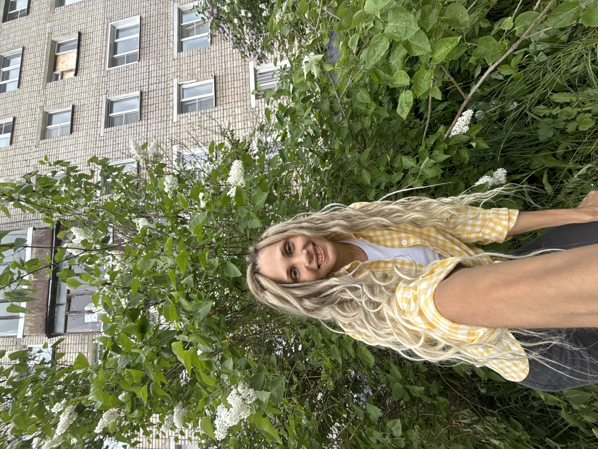 A woman with long, blonde, braided hair is smiling and looking at the camera while standing outdoors next to a flowering bush. She is wearing a white t-shirt and a yellow plaid overshirt, with dark pants. The background includes a multi-story building with numerous windows. The location appears to be an urban green space. Although it is likely taken in a city, the environment doesn’t provide enough detail to determine it is specifically Rusanovo, Russia.