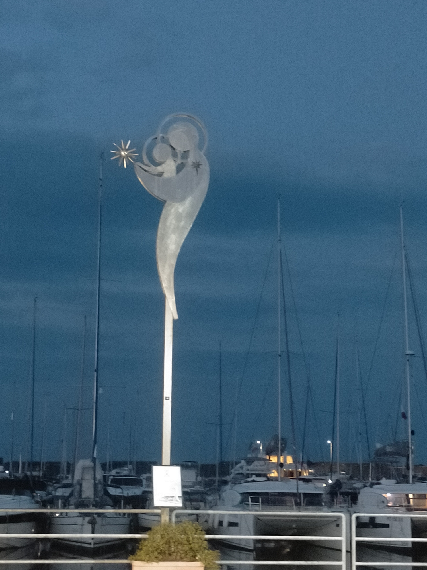 An abstract metal sculpture of a mother and child, holding a star, stands tall on a pole in a marina at dusk. The sculpture is silver and appears to be made of brushed metal. The mother's head is tilted down towards the child, who is reaching for the star. The sculpture is set against a dark blue, cloudy sky. In the background, numerous sailboats are docked, their masts forming a vertical pattern against the sky. Some of the boats have their lights on, casting a warm glow. A white sign is affixed to the pole supporting the sculpture, and a small planter with green foliage is visible at the base of the pole. The overall scene is serene and atmospheric, with a touch of artistic elegance.