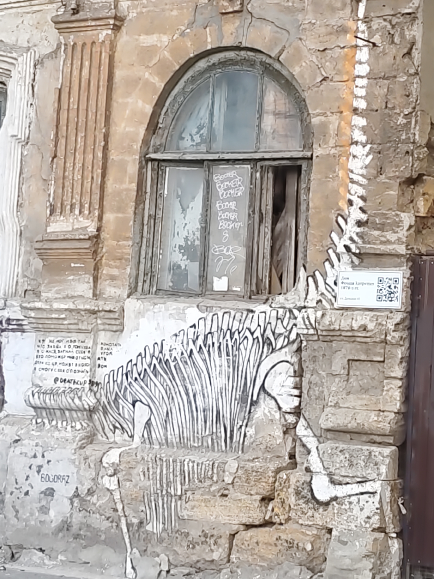 The image depicts a section of an old, weathered building in Rostov-on-Don, Russia, likely in the historical center. The architecture features a semicircular arched window with a broken pane, framed by aged, textured stone. The facade exhibits signs of decay, with peeling paint and visible cracks.
Prominently featured on the lower part of the building is a large-scale graffiti artwork of a dinosaur skeleton, rendered in black and white. The artwork is integrated into the stonework, with the ribs of the dinosaur appearing to emerge from the wall itself. Above this, there is more graffiti on the window and the wall. One section on the window shows the word "BOOMER" repeated several times, along with other illegible scribbles. On the stone to the left of the window, there is text in Russian, and a QR code with accompanying text and numbers is affixed to the wall on the right side of the dinosaur artwork, indicating the building's designation as "House of Fomina-Dmitrenko, 1970-2 pp." and "Doc. 41". Below the dinosaur artwork, the tag "BOGORAZ" is visible in large letters. The overall impression is one of urban decay and artistic intervention in a historically significant location.