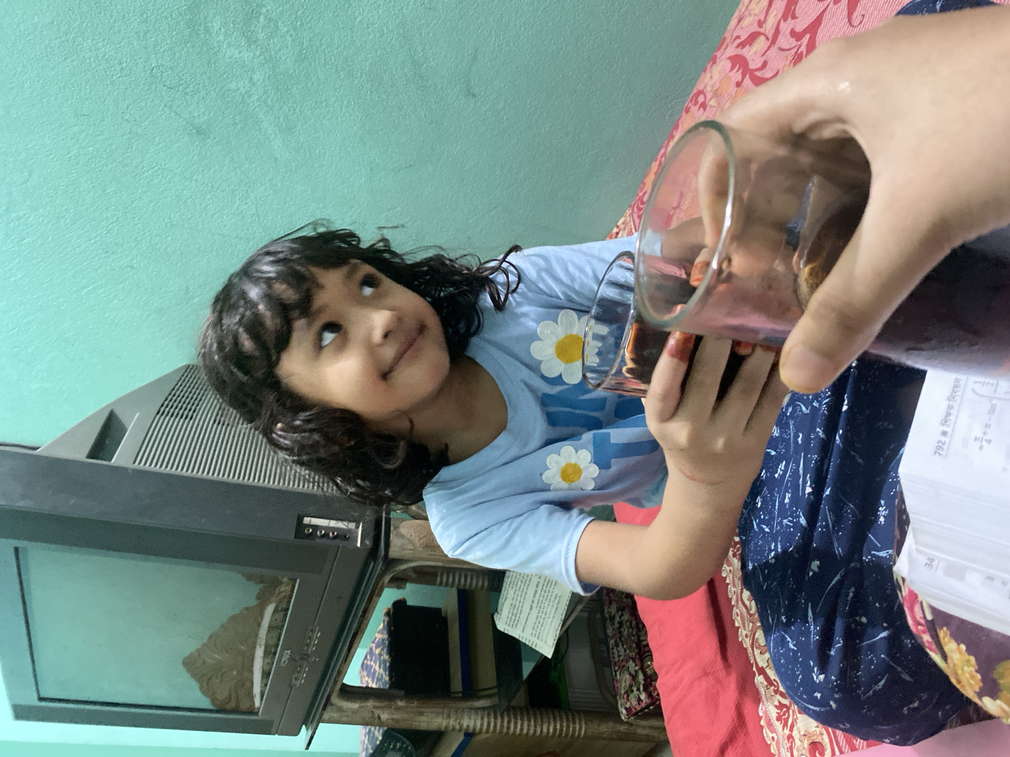 This indoor photograph captures a heartwarming moment between a young child and an unseen adult. A young girl, with curly dark hair and wearing a light blue t-shirt adorned with white daisy patterns, looks upwards with a gentle smile and an expectant expression. Her gaze is directed towards a clear glass filled with a dark red or purple liquid, which is being held by an adult's hand. The adult's right hand features intricate mehndi (henna) designs on the fingers, a cultural detail common in Dhaka, Bangladesh.
The scene takes place in what appears to be a home in Dhaka. The child is seated or lying on a bed covered with a red and pink patterned sheet, with a dark blue patterned fabric also visible, possibly another blanket or piece of clothing. To the child's left, an older model grey Cathode Ray Tube (CRT) television set is visible, resting on a wooden stand with carved details. The room's wall in the background is painted a light aqua or mint green color.
In the adult's hand, near the child, white papers with visible Bengali text are present. The text includes "792 নং" (No. 792), "বিষয়ভিত্তিক নিরসন" (Subject-wise resolution), and a partial mathematical expression " = 4+π - tan", suggesting the papers are part of a textbook or study material. The lighting in the image suggests it is daytime, with soft, indirect illumination. The overall mood is one of quiet domesticity and care, as a drink is offered to the child.