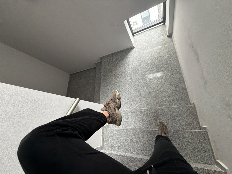 A high-angle perspective captures an individual's lower legs and feet on a modern indoor staircase. The person wears black, loose-fitting trousers and brown/taupe athletic shoes with thick, textured soles. The visible environment consists of a clean, minimalist stairwell featuring steps and landings covered in light grey speckled granite or terrazzo tiles. The walls are painted a uniform light grey. A silver-colored metal handrail runs alongside the left wall. Natural light illuminates the stairwell, emanating from a large window with a black frame positioned at the bottom right of the image, which reflects exterior building facades. The architectural style suggests a contemporary multi-story building consistent with urban construction found in Sofia, Bulgaria; however, specific identifying landmarks for a precise location within the city are not discernible from the visual evidence.
