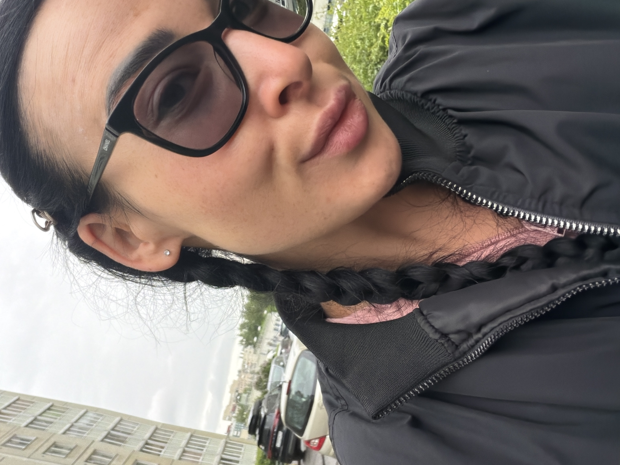 A woman is shown from the chest up. She has dark hair, styled in a braid, and is wearing sunglasses. She is wearing a black jacket and a pink shirt is visible underneath. The background appears to be a street scene. Several cars are parked along the side of the road in front of a building. The architecture suggests the location is likely in Saint Petersburg, Russia, given the style of the buildings in the background.