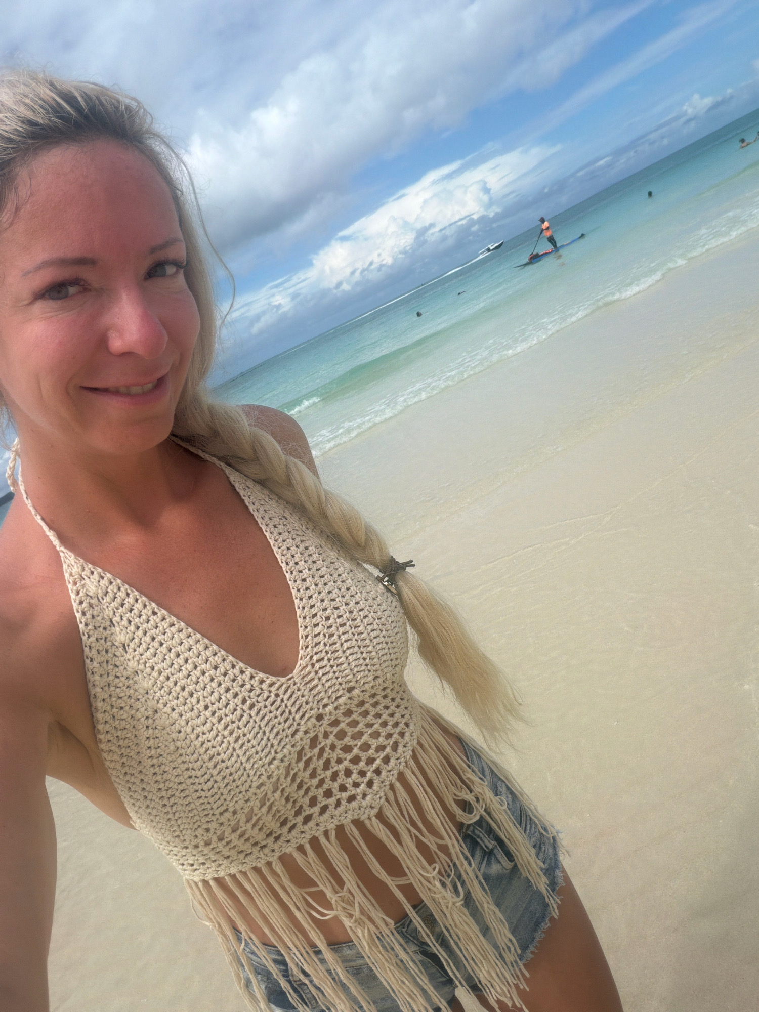A woman with blonde hair braided down her back stands on a sandy beach in Malay, Philippines, taking a selfie. She is wearing a cream-colored crochet halter top with fringe and denim shorts. The ocean, with clear turquoise water and gentle waves, stretches out behind her, meeting a bright blue sky dotted with white clouds. A person is visible paddleboarding in the distance, and a boat is also present on the water. The sand appears light-colored and wet near the water's edge.