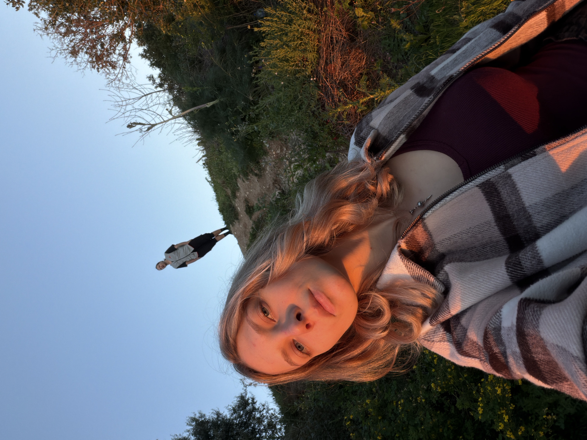 A rotated selfie photo shows a woman with blonde hair and blue eyes. She is wearing a maroon top and wrapped in a gray and white plaid blanket. The background features a green hillside with trees and vegetation. In the distance, a person is standing on the edge of a cliff, with a light blue sky behind them. The photo has warm, golden lighting, suggesting it was taken during sunset.
There is not enough information to definitively identify a location in Budva, Montenegro. More context would be required.