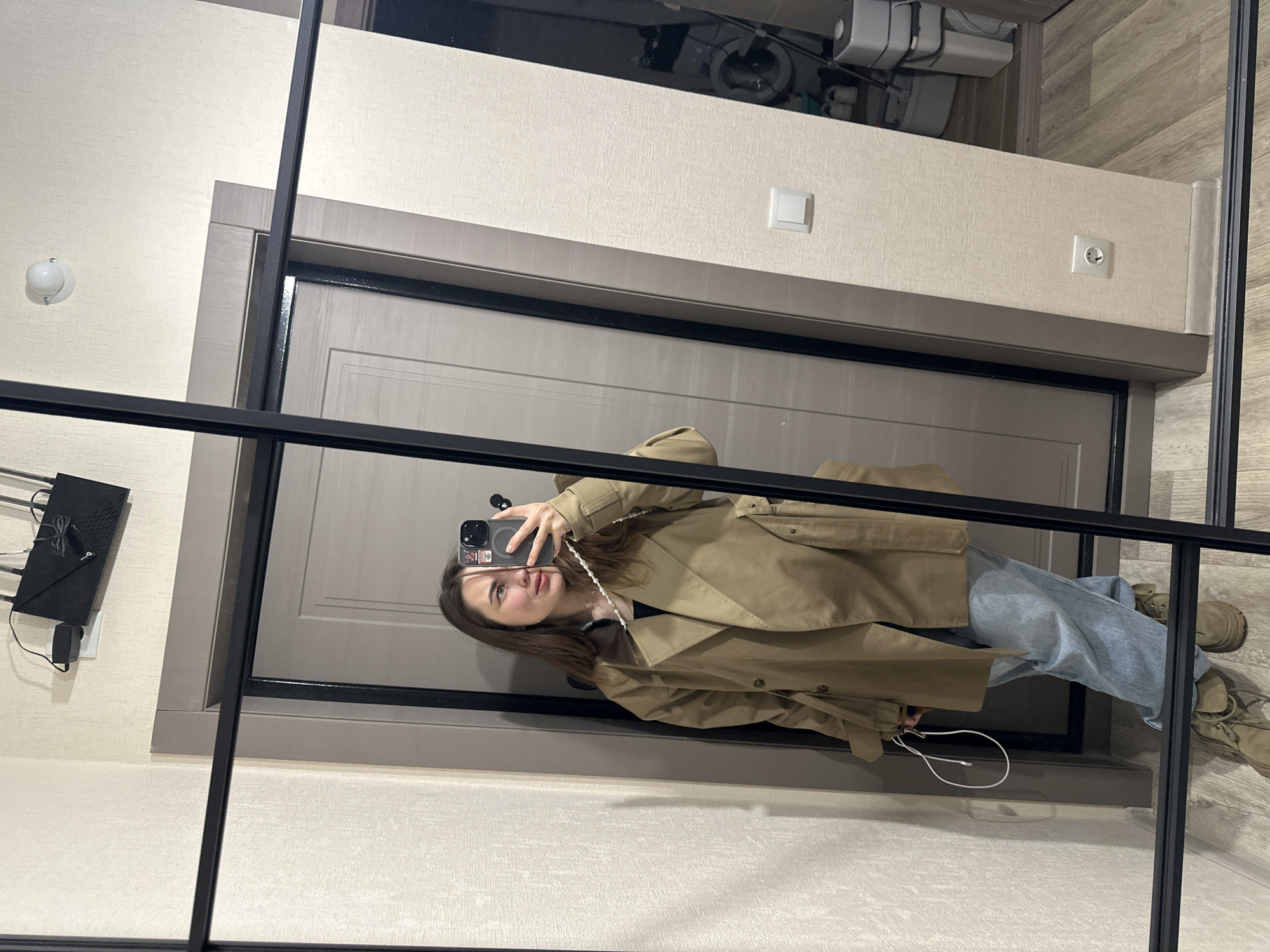 The image shows a young woman taking a mirror selfie in an indoor setting, likely a hallway or entryway in Perm, Russia. The photograph is oriented ninety degrees counter-clockwise.
The subject is a young woman with shoulder-length brown hair, looking at her smartphone as she captures her reflection. She has a neutral to slightly pouting expression. She is wearing a light brown or khaki trench coat, light blue jeans, and tan lace-up boots. She holds a black smartphone with a black case adorned with several small, indistinct stickers. A white beaded strap or chain is connected to her phone and hangs around her neck.
The background, visible as a reflection in the mirror, features light beige textured walls. To the left, a black router with multiple antennas is mounted on the wall, with visible cables and a power adapter. Further right on the wall, a white light switch and a white Type F electrical outlet are visible. The door behind the woman's reflection is a neutral gray-brown color with a simple panel design. Above the door, a shelf holds several dark and grey objects, possibly storage containers or equipment. The floor has a light wood-grain pattern. The mirror itself is framed with black vertical and horizontal bars.
The image is captured indoors under artificial lighting; therefore, the time of day and weather are not discernible. No clear, legible text is visible in the image apart from small, unidentifiable text on the smartphone case stickers.