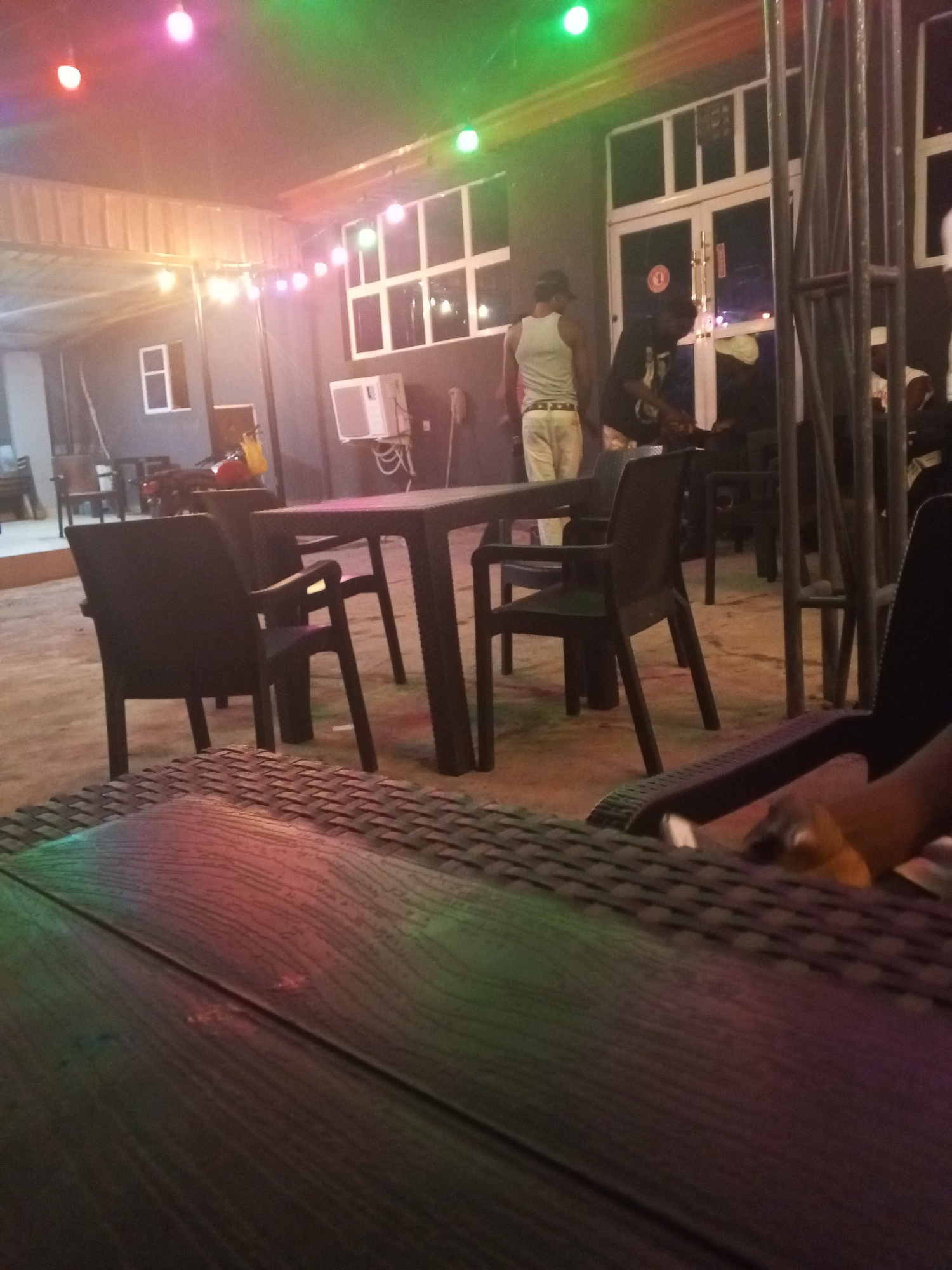 This image depicts an evening scene at an outdoor or semi-outdoor social venue, likely a bar or restaurant, located in Lokoja, Nigeria. The area is illuminated by strings of colorful lights—predominantly red, pink, green, and yellow—strung across a corrugated metal ceiling, creating a festive and dim ambiance.
In the foreground, a dark wooden table with a textured, possibly wicker, edge is visible, reflecting the vibrant colors of the overhead lights. Beyond it, several dark plastic tables and chairs are arranged across what appears to be a concrete or paved floor, set up for patrons.
Multiple individuals are present in the mid-ground and background. One man, wearing a white tank top and light-colored pants, walks away from the camera towards a white-framed double door. Another person in a dark shirt and cap stands near this door. Other figures are partially obscured by the dim lighting and metal structural elements on the right side of the frame.
The venue features dark-colored walls with several white-framed windows and the aforementioned double door, suggesting an adjacent indoor space. A white air conditioning unit is mounted on one wall below a window. A red motorcycle or scooter with a yellow bag is partially visible parked near a wall in the mid-ground. Heavy metal uprights or scaffolding are prominent on the right side, possibly supporting equipment or the structure itself.
The overall impression is one of a relaxed, casual gathering taking place after dark. No discernible text is visible within the image.