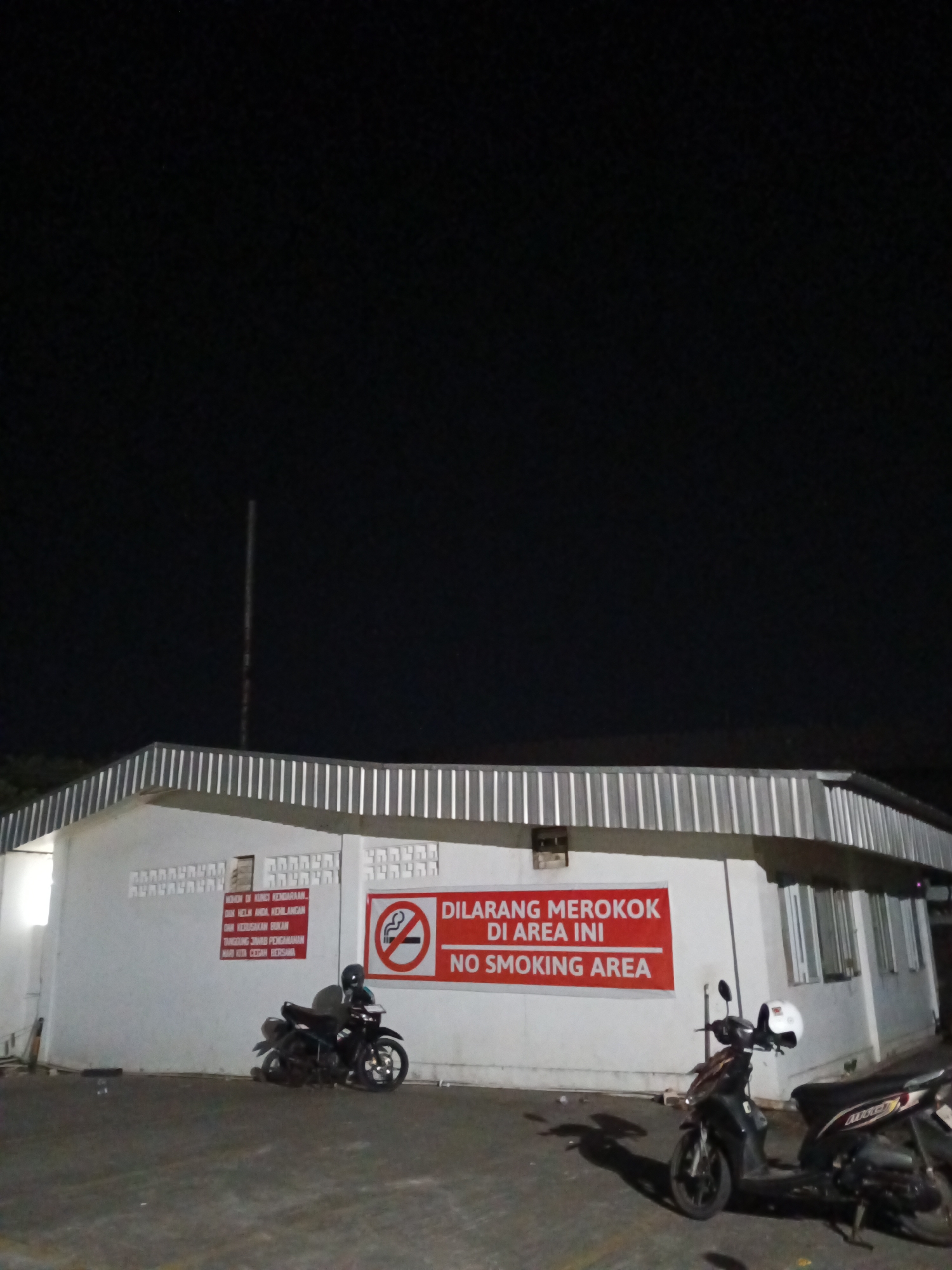 This is a low-angle, medium shot of a white, single-story building at night. The building has a corrugated metal roof and numerous windows. On the left side of the building, there is a motorcycle parked in front of a red sign with text in Indonesian and English that reads "DILARANG MEROKOK DI AREA INI NO SMOKING AREA." Another motorcycle is parked to the right of the first, with a white helmet on its handlebars. The background is dark, with the top of a tall, thin pole visible against the night sky. The scene appears to be an outdoor area, possibly a parking lot or public space. The building's facade has decorative elements that resemble brickwork. There are no people visible in the image. The overall impression is one of a functional, utilitarian building in a dark, quiet setting, with clear signage prohibiting smoking.
