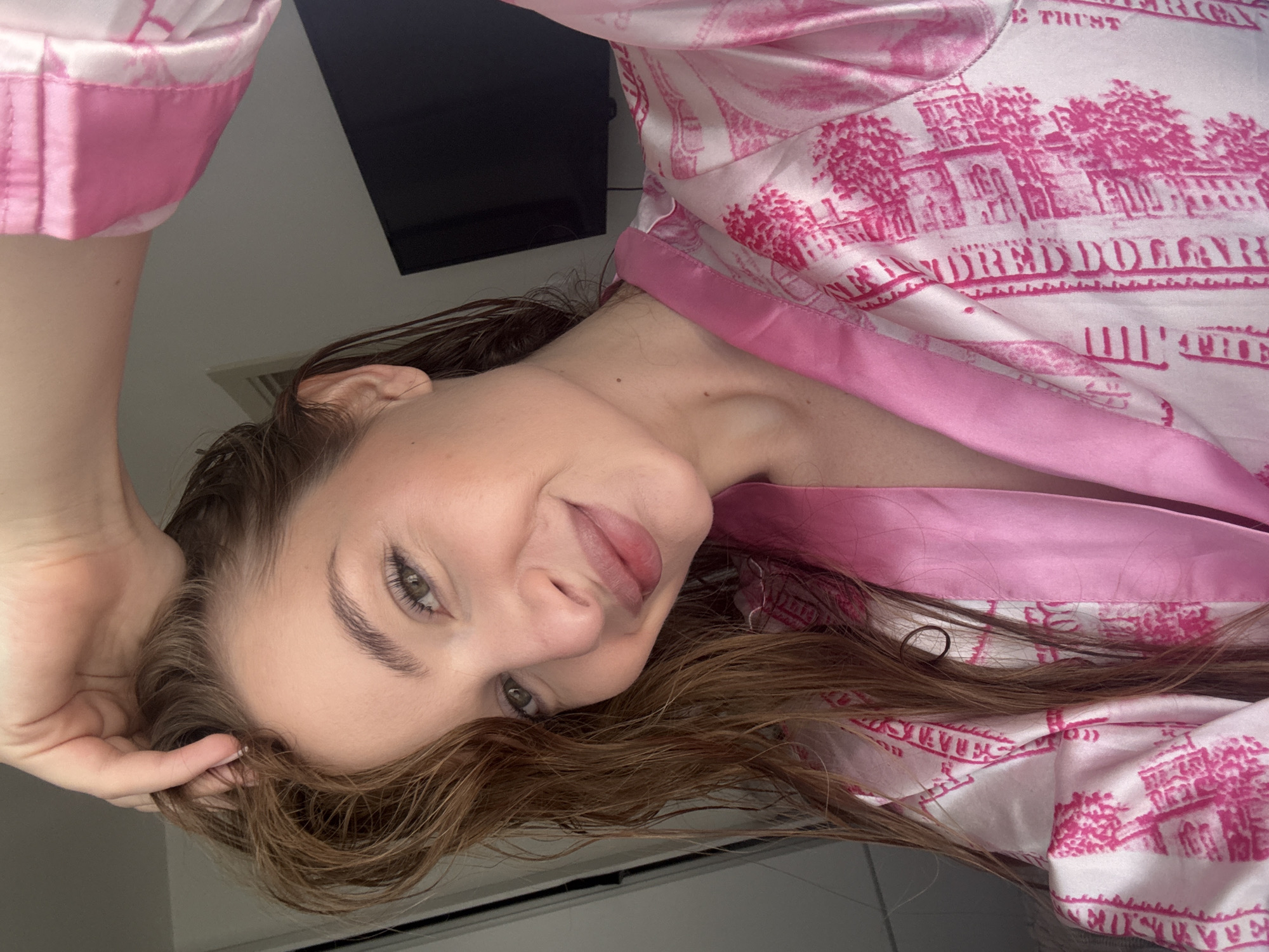 A woman with brown hair and green eyes is depicted in a close-up shot. She is wearing a pink and white robe with a pattern of the US Capitol building and the words "ONE HUNDRED DOLLARS." Her head is tilted to the side, and she is touching her hair with her left hand. A black television is visible in the background. The location is not possible to determine with the context.