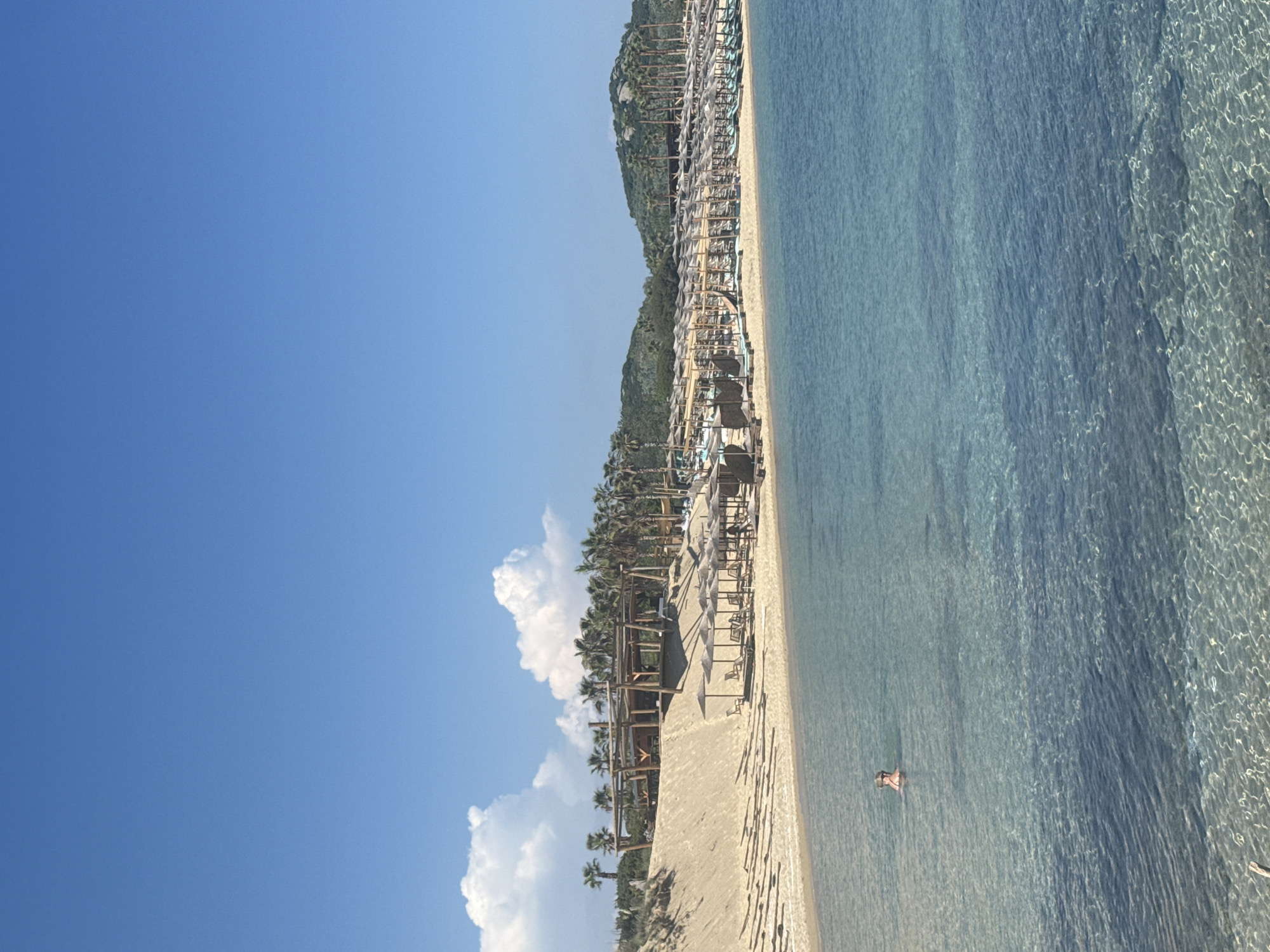The image shows a coastal scene in Nea Peramos, Greece. The view is of a beach with clear, turquoise water meeting a sandy shore. Along the beach is a structure of umbrellas and cabanas. In the distance, a low, green hill is visible along the coastline. The sky is bright blue with white cumulus clouds. A person is swimming in the water near the beach.