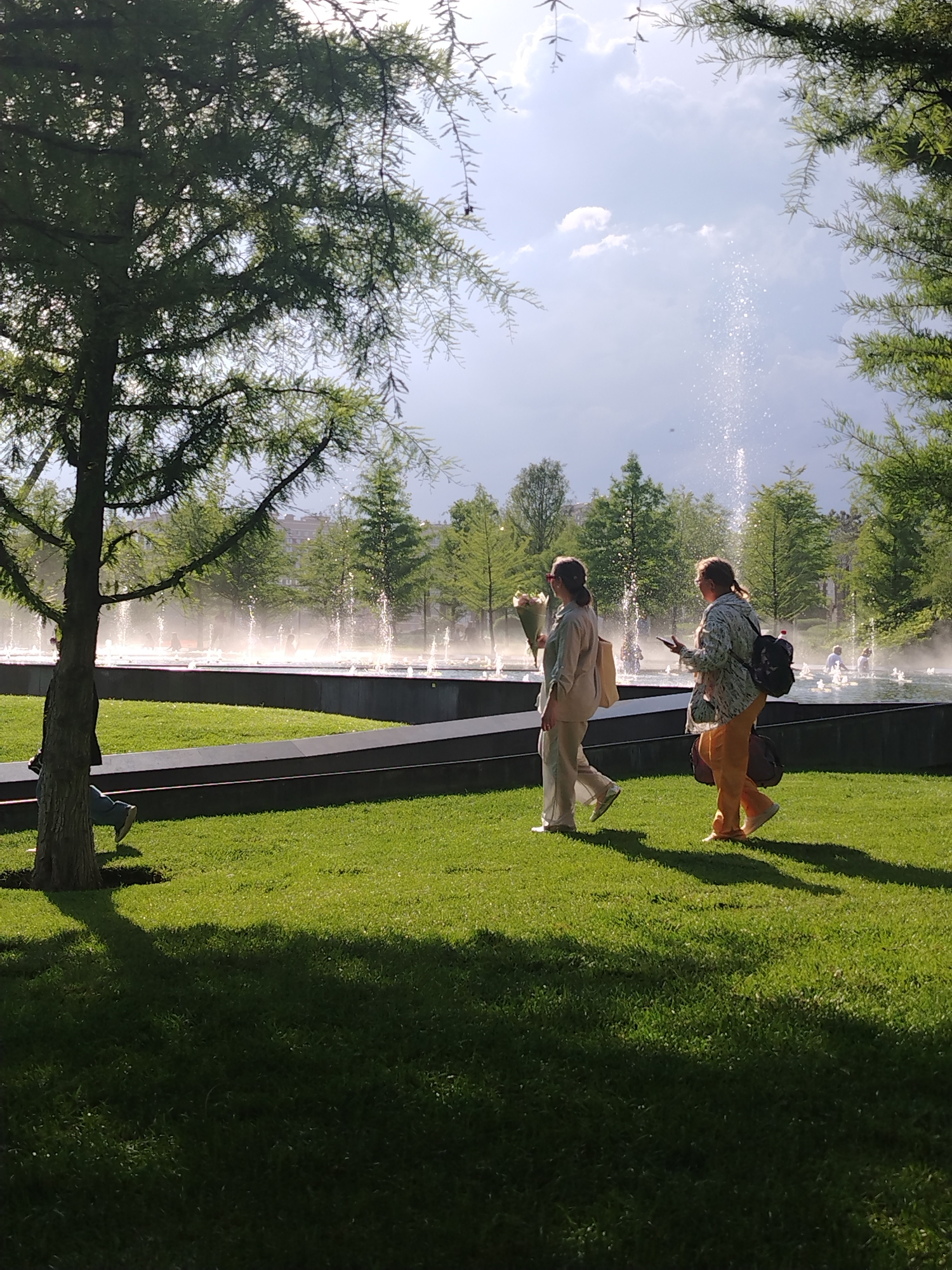 Two women walk on a grassy lawn in front of a large fountain with multiple water jets. The woman on the left wears a light-colored pantsuit and carries a light-colored tote bag. She holds a bouquet of pink and white flowers in her left hand. The woman on the right wears an olive green patterned shirt and orange pants. She has a backpack slung over her right shoulder and is holding a smartphone in her right hand. Trees with green foliage are visible behind the fountain and framing the shot on the left. The sky is blue with scattered white clouds and the sun is shining, casting shadows on the grass. In the distance, buildings can be seen through the trees. The scene appears to be in a park or public space.