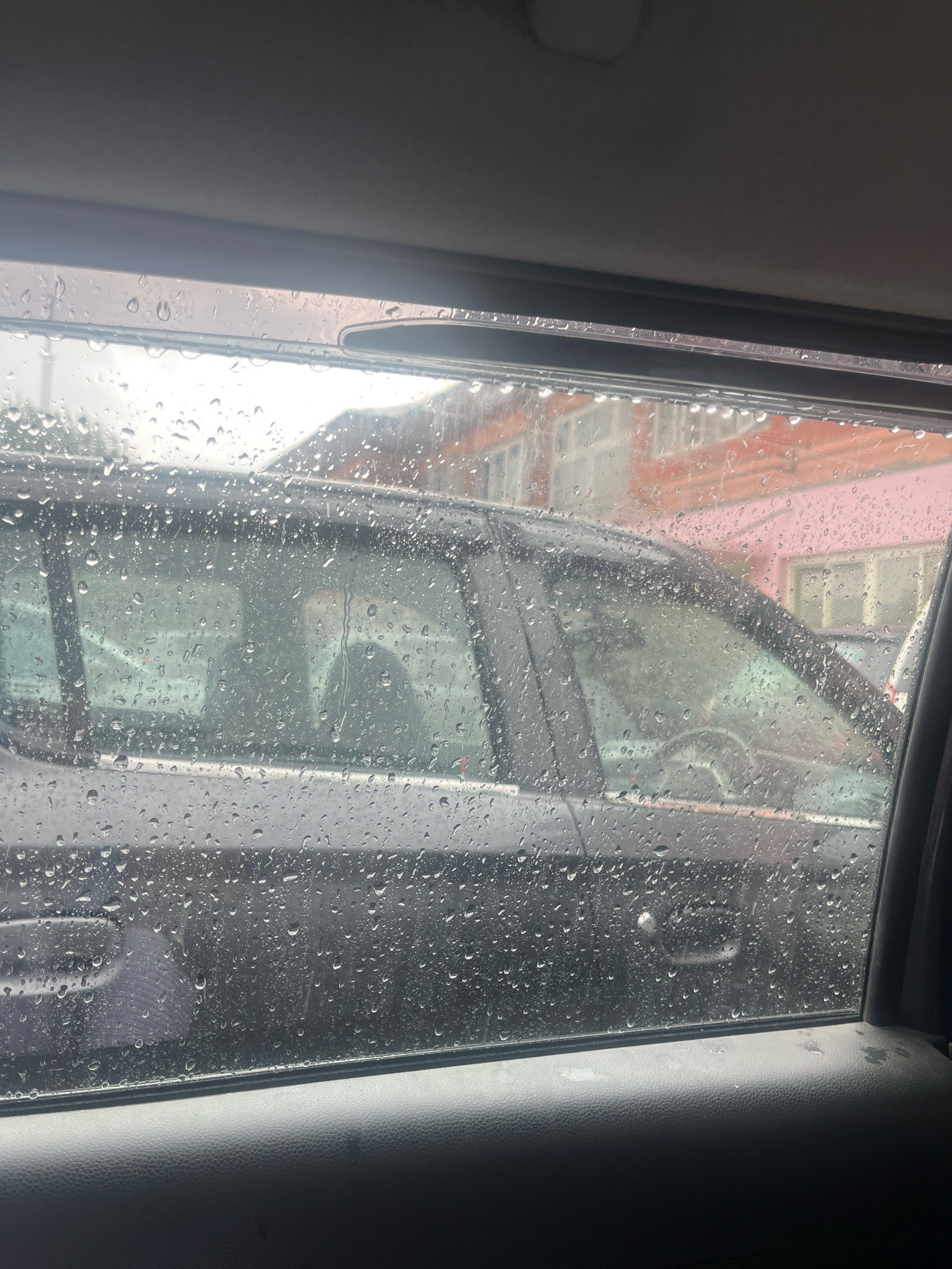 A view from inside a vehicle, looking out a rain-streaked window at another vehicle parked outdoors. The window is covered in numerous water droplets, obscuring the view of the exterior. Beyond the second vehicle, a building with pink and orange sections is visible, suggesting an urban or suburban setting. The interior of the vehicle from which the photo is taken is dark, with the textured surface of a door panel visible in the foreground. The overall impression is one of a rainy day in Kupwara, India.