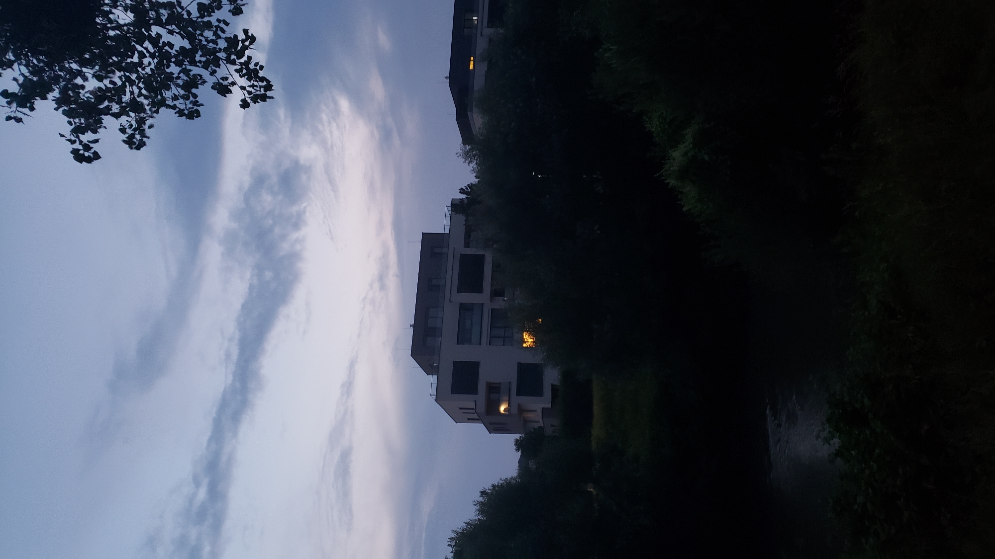 The image depicts a modern apartment building situated in a dark, natural environment, likely near a body of water, in Prešov, Slovakia. The building is white with dark-framed windows and balconies, and some interior lights are visible, casting a warm glow. The foreground and surrounding areas are heavily shadowed by dense foliage, including trees and bushes, obscuring the ground level and a portion of what appears to be a river or stream on the left side. The sky is a dusky blue with wispy clouds, suggesting it is either dawn or dusk. A tree branch with leaves is visible in the upper right corner, also silhouetted against the sky. No people are visible in the scene.