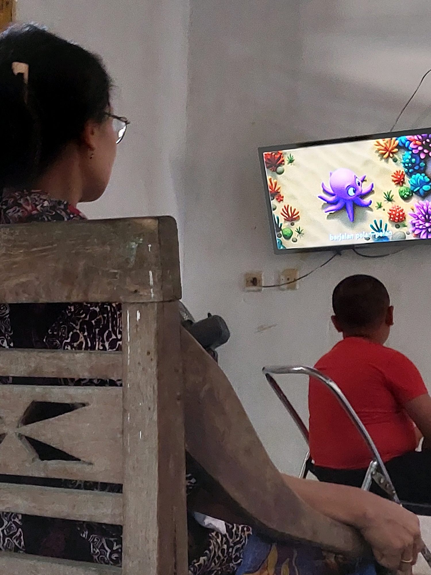 A medium shot captures two individuals seated, facing a television screen displaying animated marine life, an octopus being the prominent subject. The scene appears to be an indoor domestic setting, possibly a living room, with a white wall as the backdrop.
In the foreground, to the left, a person with dark hair tied back and wearing glasses is seated in a wooden chair, their back to the camera. They are dressed in a patterned garment. The wooden chair has an intricately carved design on its backrest.
To the right, a young child, viewed from the back, is seated in a metal-framed chair, wearing a red t-shirt and dark pants. The child is also looking towards the television.
The television, mounted on the wall, shows a vibrant cartoon scene of an underwater environment with coral reefs and a purple octopus. Text in a language other than English is visible at the bottom of the screen. Electrical outlets are present on the wall behind the child. A thin black wire runs from the television to a point on the wall.
The general environment is uncluttered, suggesting a simple living space. The lighting appears to be artificial, contributing to a slightly dim atmosphere. The image lacks any specific indicators that would pinpoint the exact location within Sarangan, Indonesia, beyond it being a domestic interior.