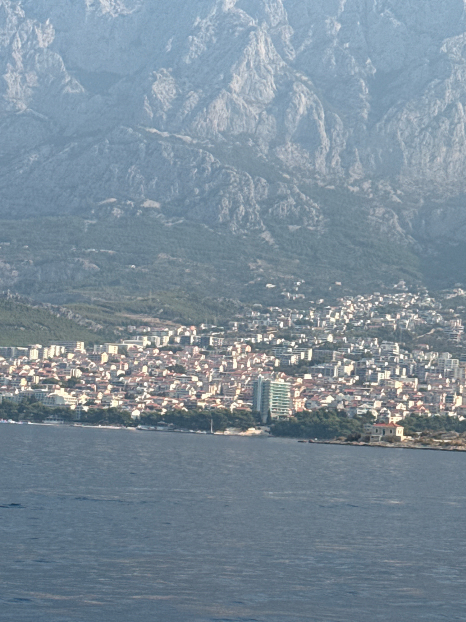 The image shows an aerial view of the city of Makarska, Croatia, situated on the coast with mountains in the background. The city is characterized by densely packed buildings with red-tiled roofs, interspersed with some taller, modern structures. A distinct, tall, multi-story building with a light-colored facade and vertical accents stands out among the lower buildings. The city is bordered by lush green trees along the coastline. The sea in the foreground is a deep blue with subtle ripples on its surface, indicating a calm body of water. The overall scene captures a sunlit coastal city nestled against a dramatic mountainous landscape.
