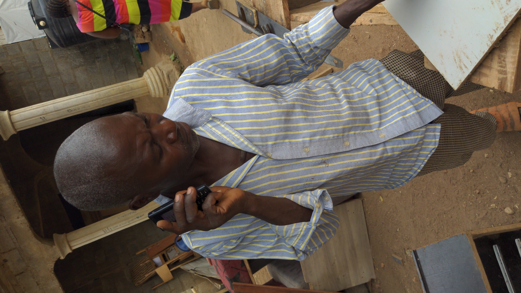 An overhead, rotated photograph captures a man interacting with a feature phone in what appears to be an outdoor or semi-outdoor workshop environment in Masaka, Nigeria.
The main subject is a man with dark skin, closely cropped greying hair, and a slight beard. He is wearing a long-sleeved, blue and yellow striped shirt, partially unbuttoned, and brown/black checkered trousers. He holds a black feature phone with a physical keypad in his right hand, his eyes squinted as if looking at the screen or into sunlight.
In the background, another person, with their back to the camera, wears a vibrant striped shirt (pink, orange, yellow, black) and dark pants, carrying a black bag. Further back, a leg with an orange-brown shoe or sandal is visible.
The setting includes bare, dusty ground and several architectural elements like two light-colored, ornate pillars. There are also various wooden materials, including planks and what appears to be a saw, suggesting a carpentry or construction area. A white rectangular panel, possibly a board, rests on wooden blocks.
The scene appears to be taking place during the daytime, under bright, sunny conditions. On one of the pillars, faint handwritten text is visible, including what looks like "OARAA" and a "Q" or "O".