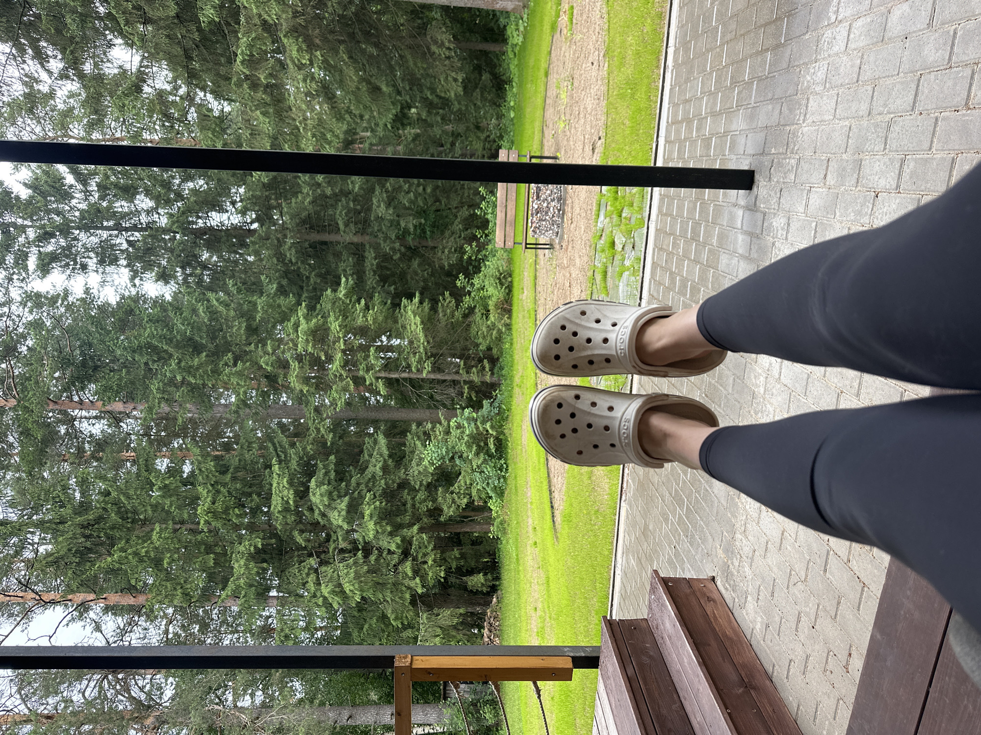 The image depicts a person's legs, clad in black leggings and white Crocs, resting on a covered patio. The patio floor is paved with grey interlocking bricks. To the left, wooden steps lead up to the patio. Beyond the patio, a lush green lawn stretches towards a dense forest of tall pine trees. In the distance, a wooden bench sits near a circular fire pit area with gravel and stones. The scene suggests a relaxed outdoor setting, possibly a backyard or a cottage in a wooded area. The overall impression is one of tranquility and nature.