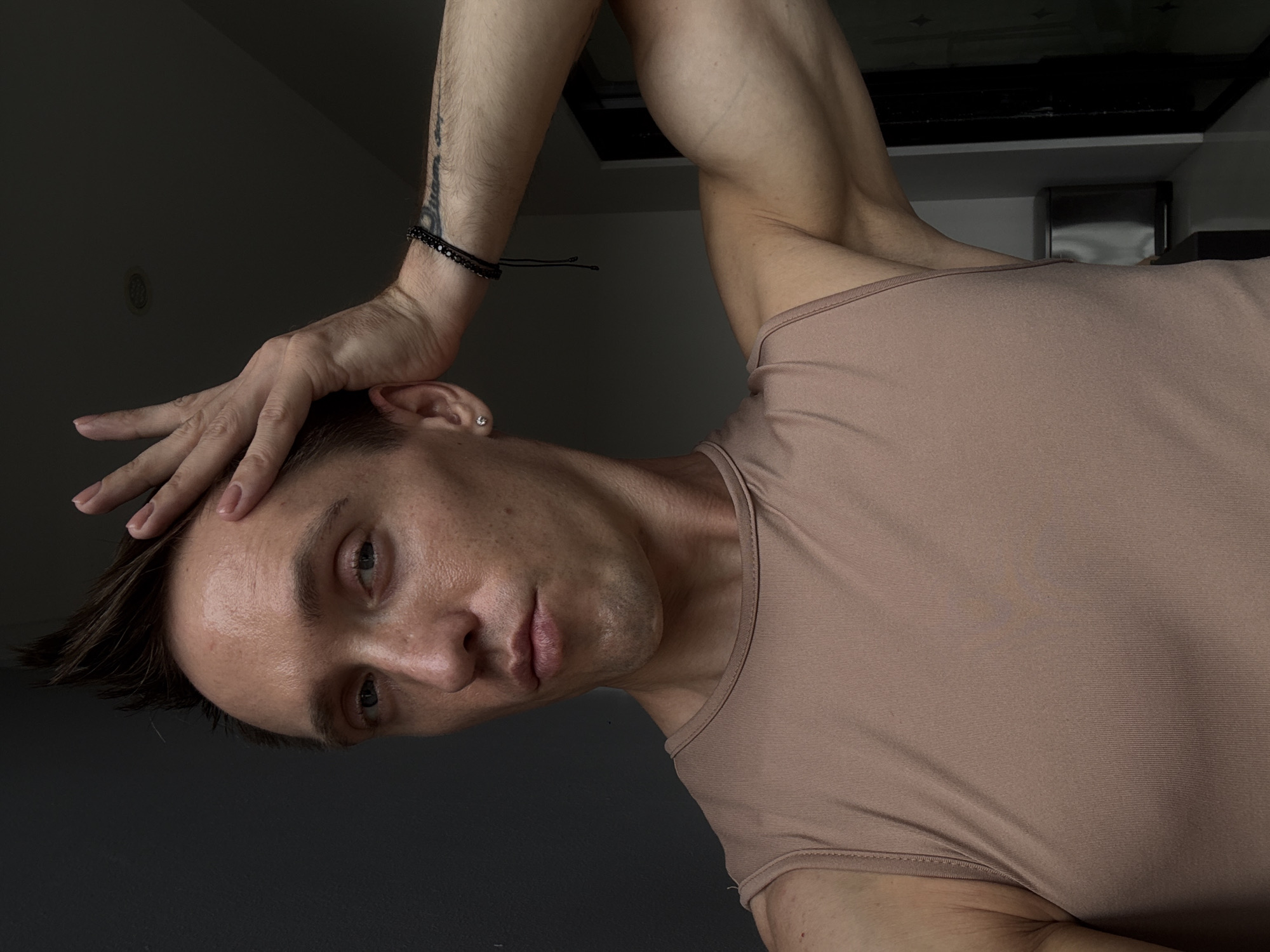 A person is posing with their hand on their head and their arm flexed. They are wearing a tan, sleeveless top. The background is dark, with a gray wall on the left and a doorway on the right. A trash can is visible to the right of the person. The lighting is dim, creating shadows on the person's face and body. The image appears to have been taken indoors.