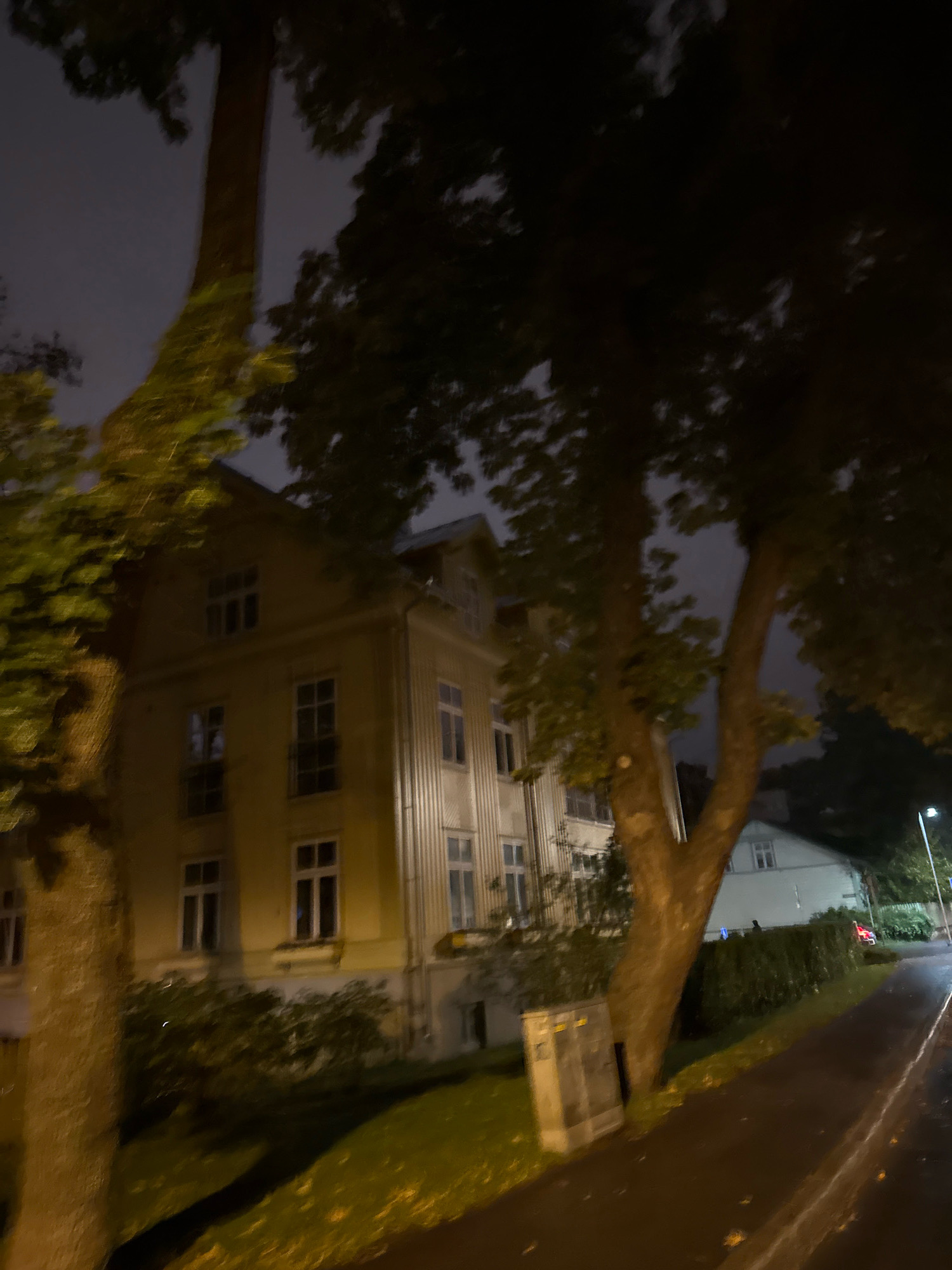 An exterior night shot captures a building and street in Tallinn, Estonia. The building, painted a pale yellow, has multiple windows on each of its visible floors. Several trees with dark leaves partially obscure the building. A paved walkway runs along the right side of the image, and a street light illuminates the scene. A grey utility box stands near the base of a tree on the left. In the background, a lighter-colored building is visible behind a hedge. The sky is dark and overcast.
