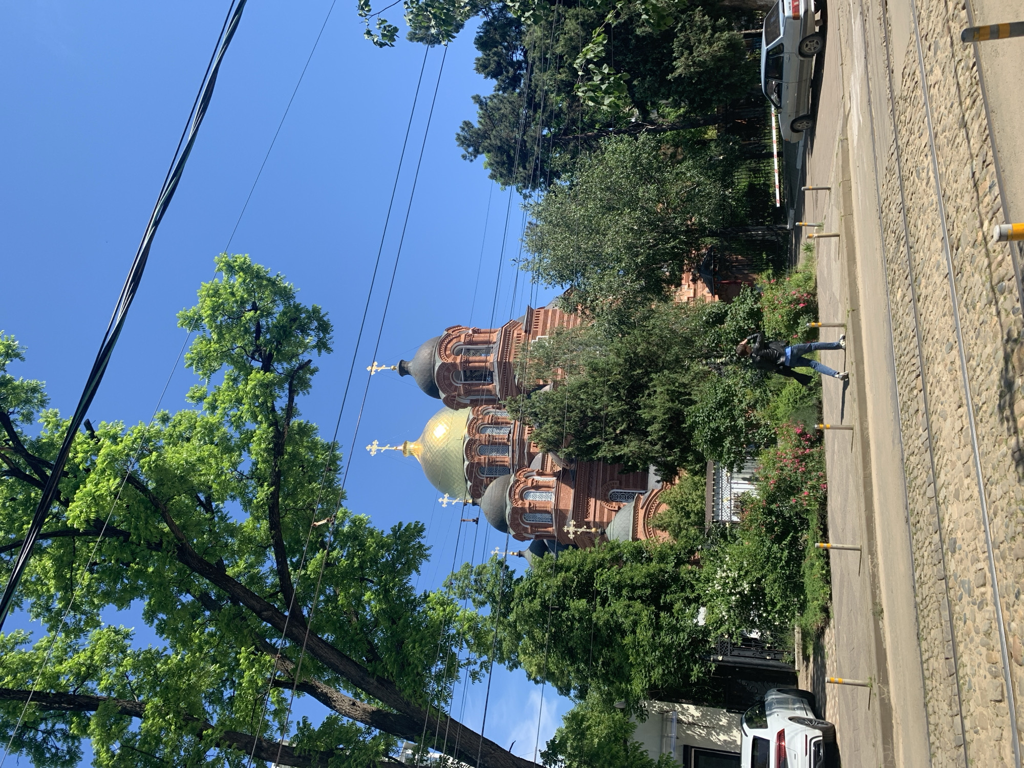 The image captures a vibrant street scene dominated by a large, ornate brick church under a clear blue sky. The church features multiple domes; at least two golden ones with crosses prominently displayed, and several smaller, darker domes. Its facade is a rich reddish-brown brick, adorned with intricate architectural details and arched windows.
Numerous lush green trees frame the church, their foliage partially obscuring some of its features. Overhead, a complex network of utility wires crisscrosses the expansive blue sky.
To the right of the image, a paved sidewalk runs alongside what appear to be tram tracks embedded in cobblestones. Several light-colored bollards line the edge of the sidewalk. A man, dressed in a dark jacket and jeans, is visible walking on the sidewalk. Further down the street, two cars are present: a light-colored sedan parked near the sidewalk and a white SUV partially visible. The scene is brightly lit by the sun, indicating midday or early afternoon, with distinct shadows cast. No visible text is identifiable in the image.