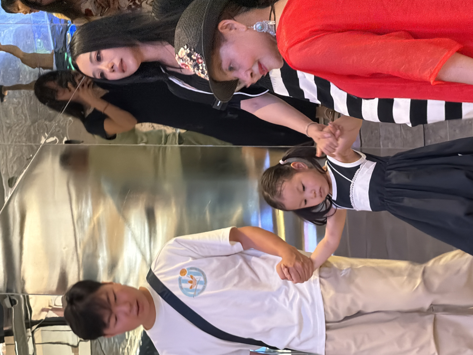 An outdoor, mid-shot photograph shows four people on a street in Yuzhong District, China, with a mirrored wall in the background. On the left is a young Asian male wearing a white t-shirt with a blue and orange logo and light-colored pants, carrying a black strap across his chest. To his right, a young Asian girl in a black and white dress holds his hand. Behind the girl is another Asian woman in a black and white striped dress and a black straw hat adorned with flowers. To the right of her, a woman in a red top and black pants is visible. The lighting is bright, casting reflections on the mirrored wall.