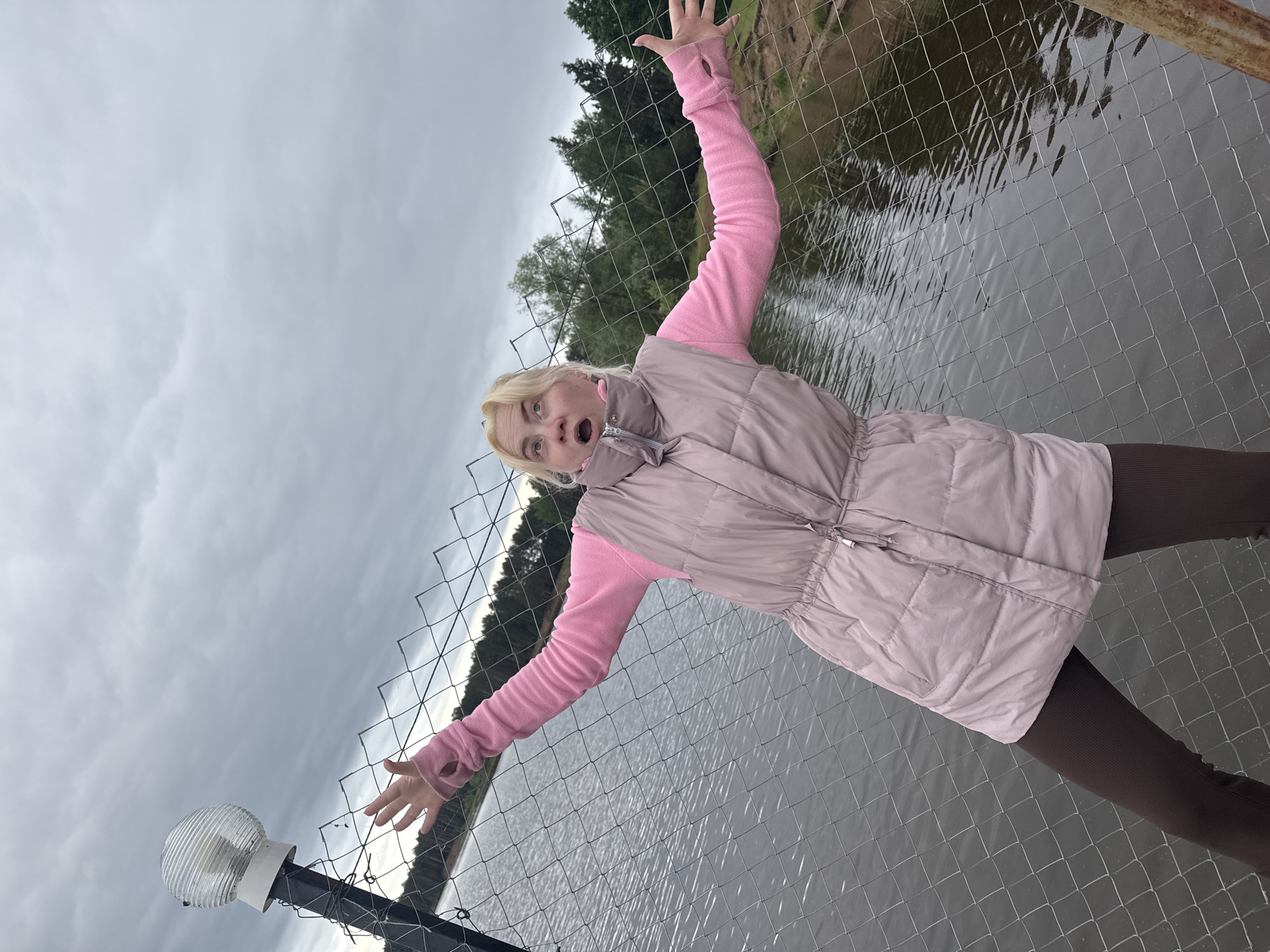 A woman with light blonde hair, wearing a pink long-sleeved shirt and a light pink vest, stands beside a wire fence with water visible behind it. She has her arms outstretched, hands on the fence, and an open-mouthed expression. A light fixture is attached to a pole near the fence. The sky is overcast. The location is identified as Chaikovsky, Russia.