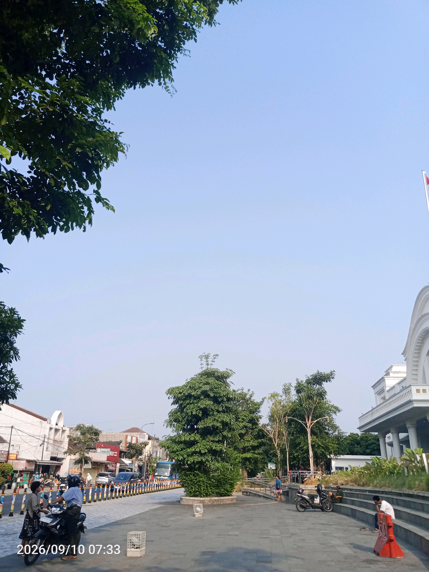 A daytime, outdoor, eye-level shot depicts a public area with a paved walkway and a street in the city of Surakarta, Indonesia. The sky is clear and blue. To the left, large green trees with lush foliage overhang the upper portion of the image.
In the foreground, a paved plaza area is visible. Two people are standing next to a parked motorcycle on the left side of the plaza. Further down the plaza, a person is seated on a concrete step, leaning their head on their hand, with a red traffic cone nearby. Another person is seated on a step further back, next to a parked motorcycle.
Beyond the plaza, a street is lined with buildings and trees. Several vehicles, including cars and a bus, are present on the street. Colorful bollards separate the street from the walkway. A large, white building with classical architectural elements, including columns and arches, is visible on the right side of the image. A flagpole with a red and white flag is at the top of the building. The timestamp "2026/09/10 07:33" is overlaid on the lower-left corner of the image.
