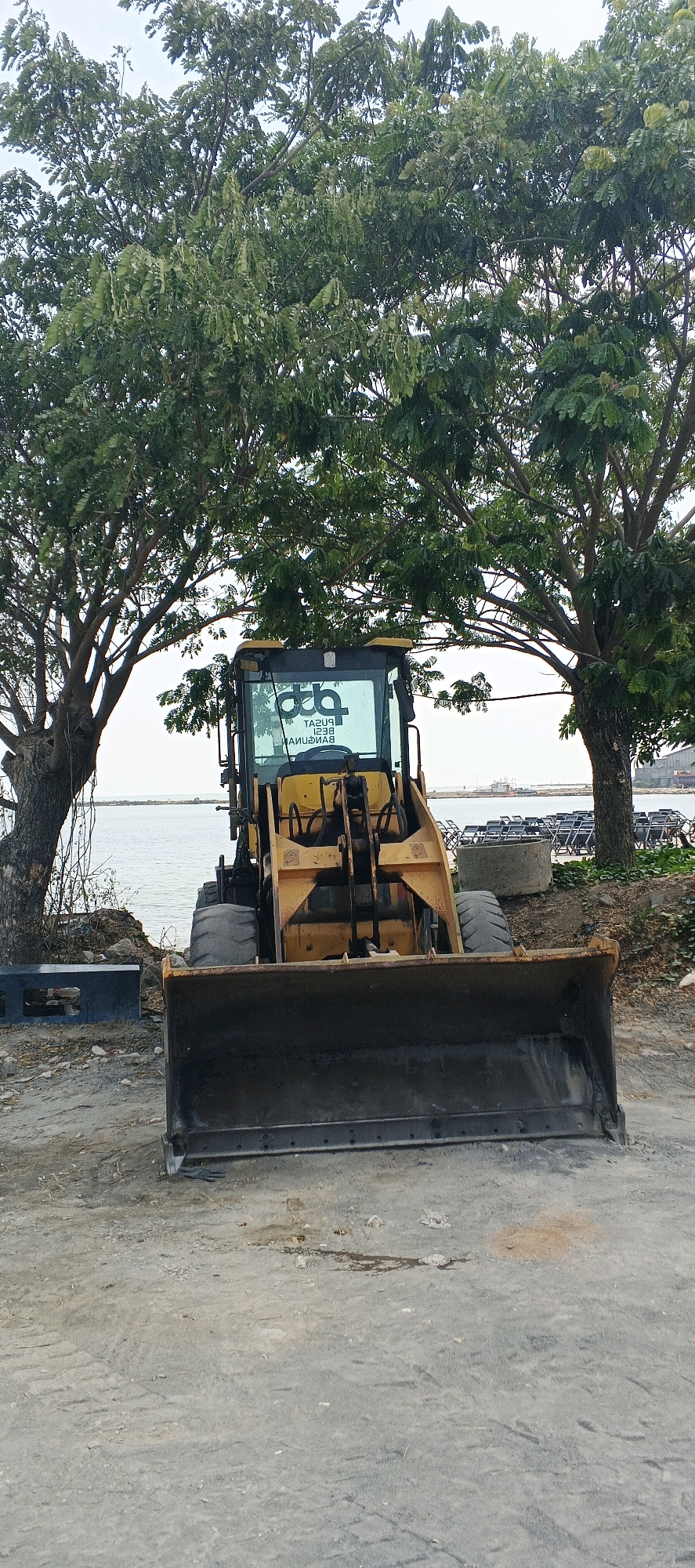 An unoccupied yellow front-end loader is parked on a dirt and gravel lot near a body of water. Two large trees with dense green foliage frame the loader. The loader has a large black bucket in front and a cab with a logo on the windshield. In the background, there are stacks of chairs, concrete pipes, and boats on the water. The sky is overcast.