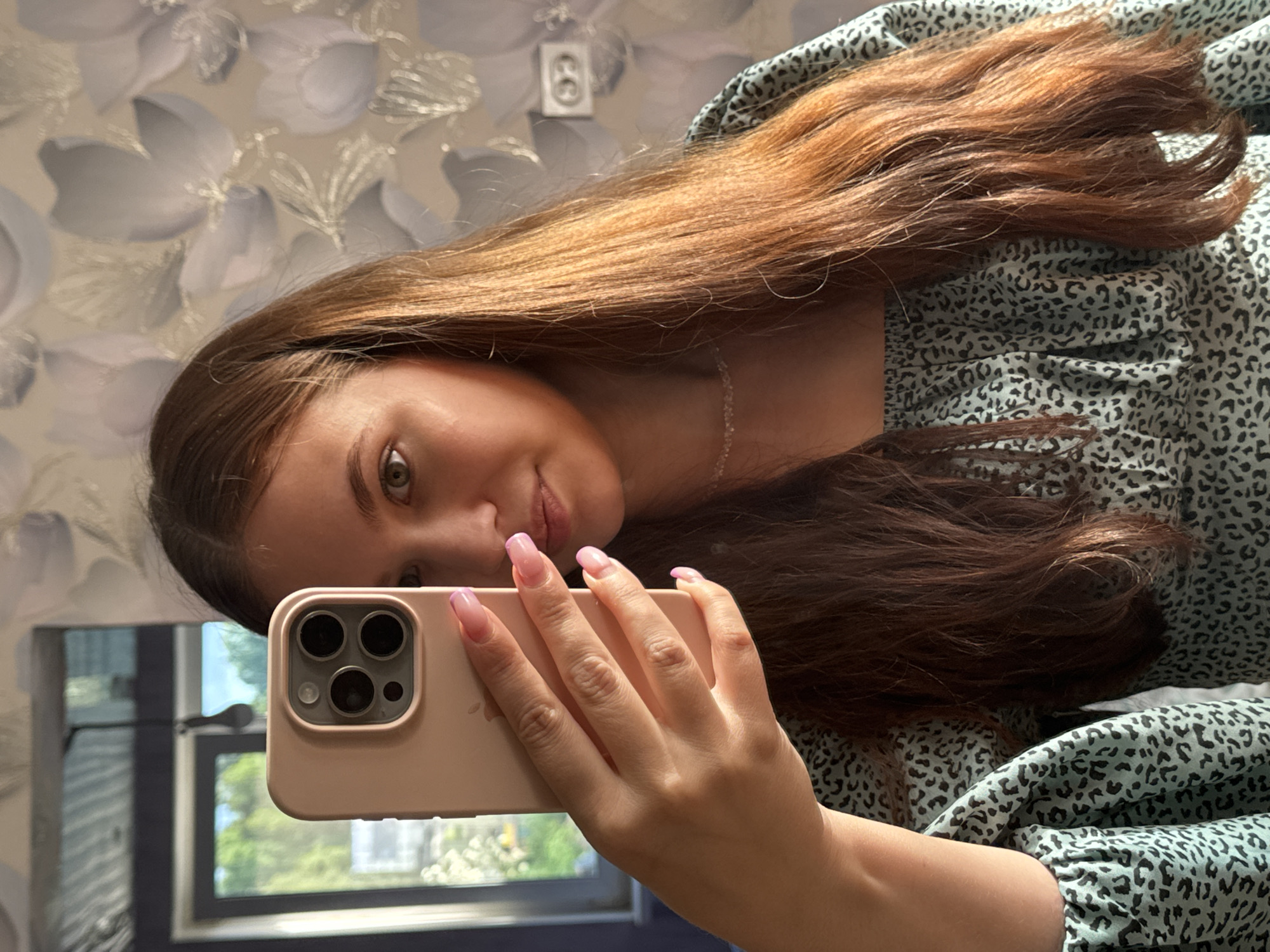 A young woman with long, reddish-brown hair is taking a mirror selfie in a room. She is holding a light pink smartphone up to the mirror with her left hand. Her nails are painted with a light pink French manicure. She is wearing a light blue, off-the-shoulder top with a black leopard print pattern. A delicate, thin necklace is visible around her neck. The background of the room includes a window with greenery visible outside, and patterned wallpaper on the wall. The overall scene suggests an indoor, domestic setting. There is no indication of the city of Nizhnyaya Gar', Russia within the image.