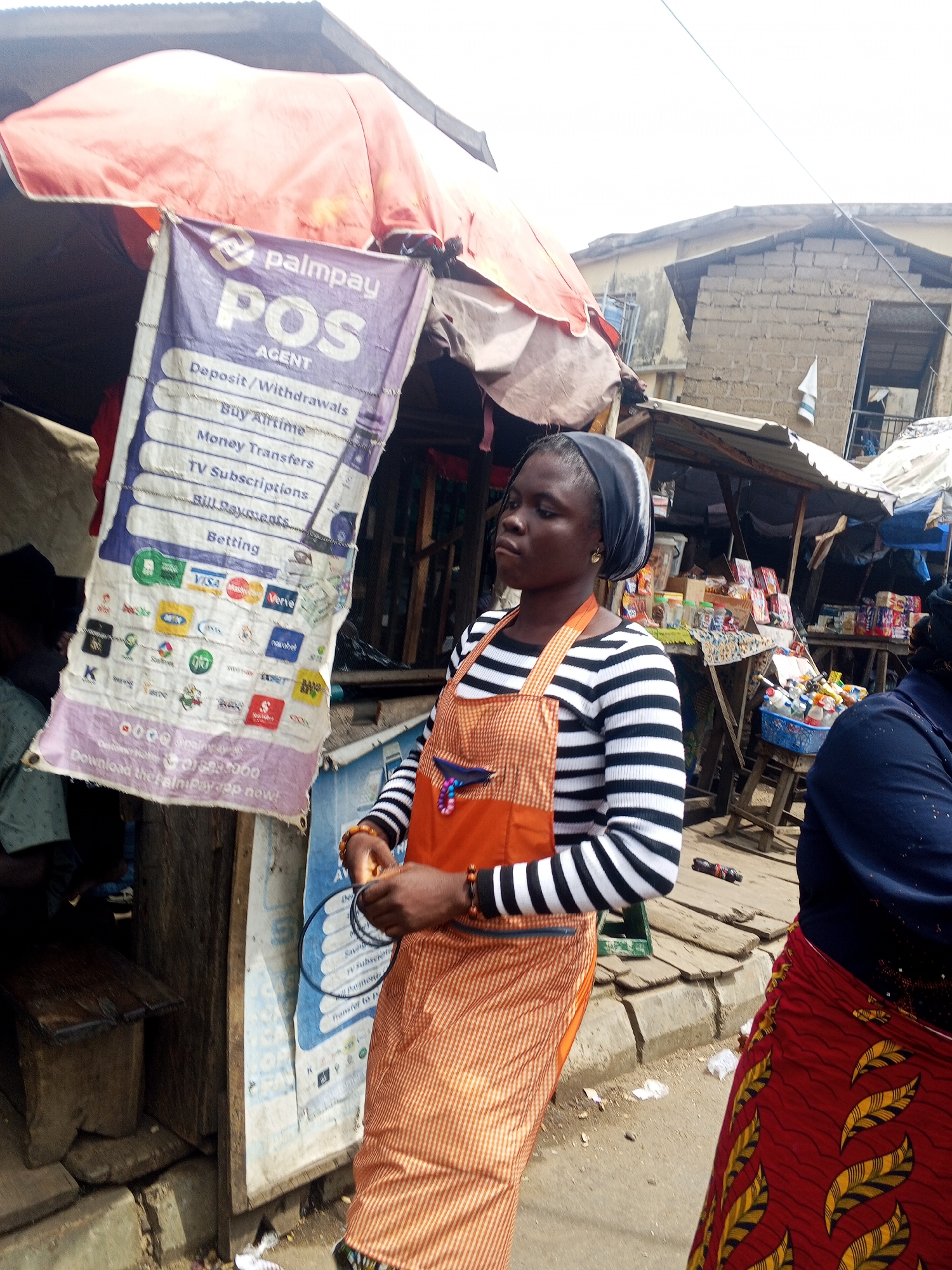 A medium shot captures a woman standing outdoors in a market in Okene, Nigeria. She is positioned in front of a stall adorned with a purple and white sign that reads "palmpay POS AGENT" and lists various services. The woman wears a black and white striped long-sleeved shirt and an orange apron with vertical stripes. She has a dark headscarf and is looking to her left with a neutral expression. Her hands are in the foreground, holding dark circular objects. To her right, another person, mostly obscured, wears a navy blue top and a red and yellow patterned skirt. The market appears to be bustling, with stalls visible in the background, displaying goods and materials. The ground is paved and appears to be a dirt or gravel surface. The sky is overcast.