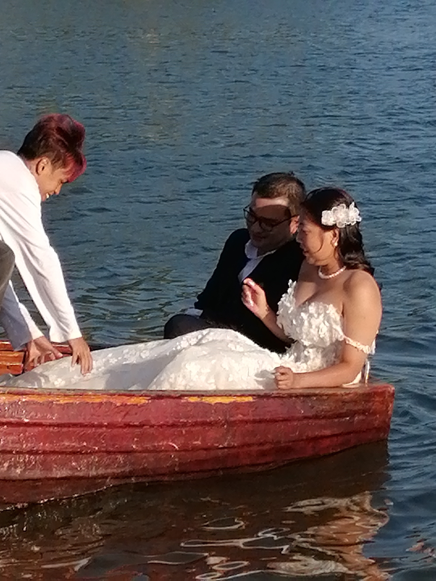 In Da Lat, Vietnam, a wedding photoshoot is taking place in a small, red rowboat on a body of water. A man in a white long-sleeved shirt and grey pants is leaning into the boat from the left, reaching down towards a woman seated in the boat. The woman is wearing a white strapless wedding dress adorned with floral appliques and a pearl necklace. She has a white floral hair accessory in her dark hair. Beside her, a man in a black suit jacket and glasses is seated. The boat is red and appears worn, with visible layers of paint. The water is a deep blue with gentle ripples.