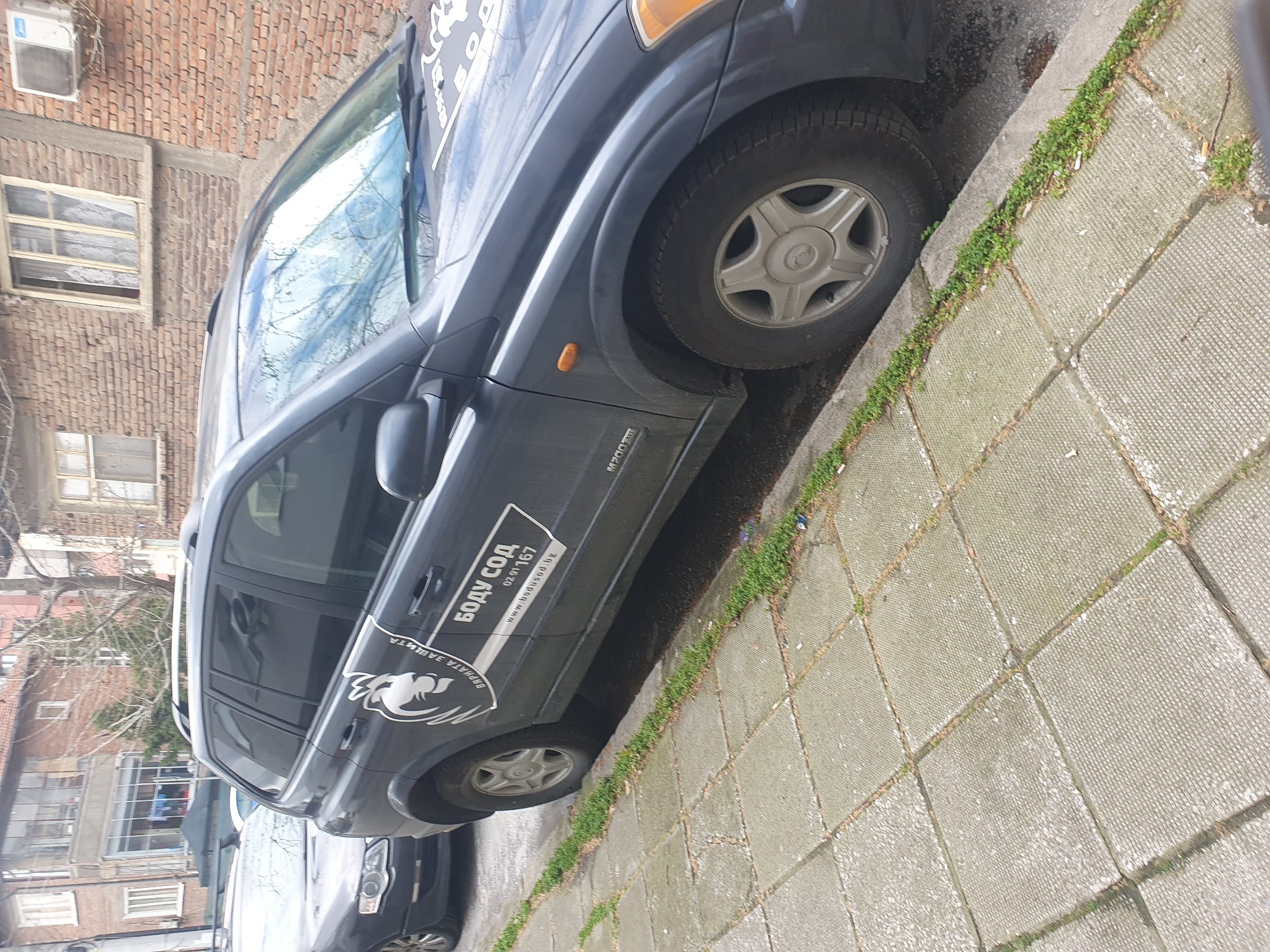 The image shows a gray SsangYong Kyron SUV parked partially on a sidewalk in Plovdiv, Bulgaria. The SUV has a company logo and contact information on the side. The sidewalk is paved with square concrete slabs, with grass growing in the joints. In the background, there is a brick building with several windows and an air conditioning unit. Other vehicles are parked nearby, and bare tree branches are visible.