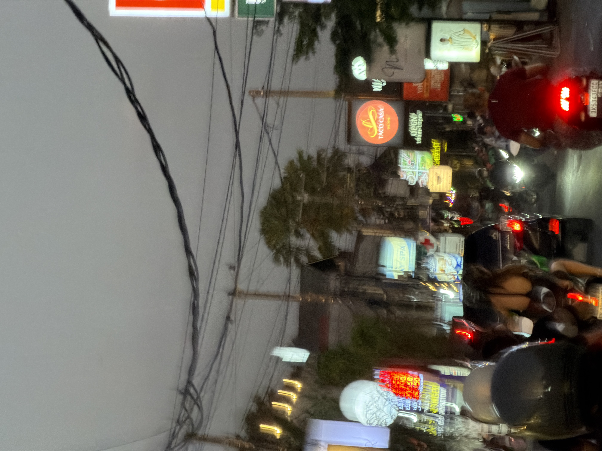 An evening street scene in North Kuta, Bali, Indonesia, is captured with significant motion blur, indicating movement or low-light conditions. The sky is dark grey and obscured by a dense network of entangled overhead electrical cables supported by utility poles that crisscross the upper frame. Below, a busy commercial street is lined with multi-story buildings, interspersed with palm trees and dense foliage.
Numerous brightly illuminated neon and LED signs advertise various businesses. A prominent round, orange sign for "Taco Casa EST. 2010" is visible in the mid-right. To its right, a partially visible red and green sign suggests a "7-Eleven." Below Taco Casa, an "OPEN 10AM-10PM" sign is displayed. Other visible signage includes a blue sign for "Y SPA," a stylized "N" logo, a rectangular sign displaying a striped shirt, and various other colorful, blurry signs, some appearing to advertise currency exchange services.
The street is densely populated with traffic, primarily motorcycles. In the foreground, a person wearing a black helmet is partially visible on the left. In the mid-ground on the left, a person with long dark hair, wearing a dark top, is riding as a passenger on a motorcycle, facing away from the viewer. On the right, a person in a red shirt and black helmet is riding a motorcycle, displaying an Indonesian license plate "DKS 343 FCC." Red brake lights and white headlights from multiple vehicles illuminate various parts of the scene, confirming active vehicle movement along the street.