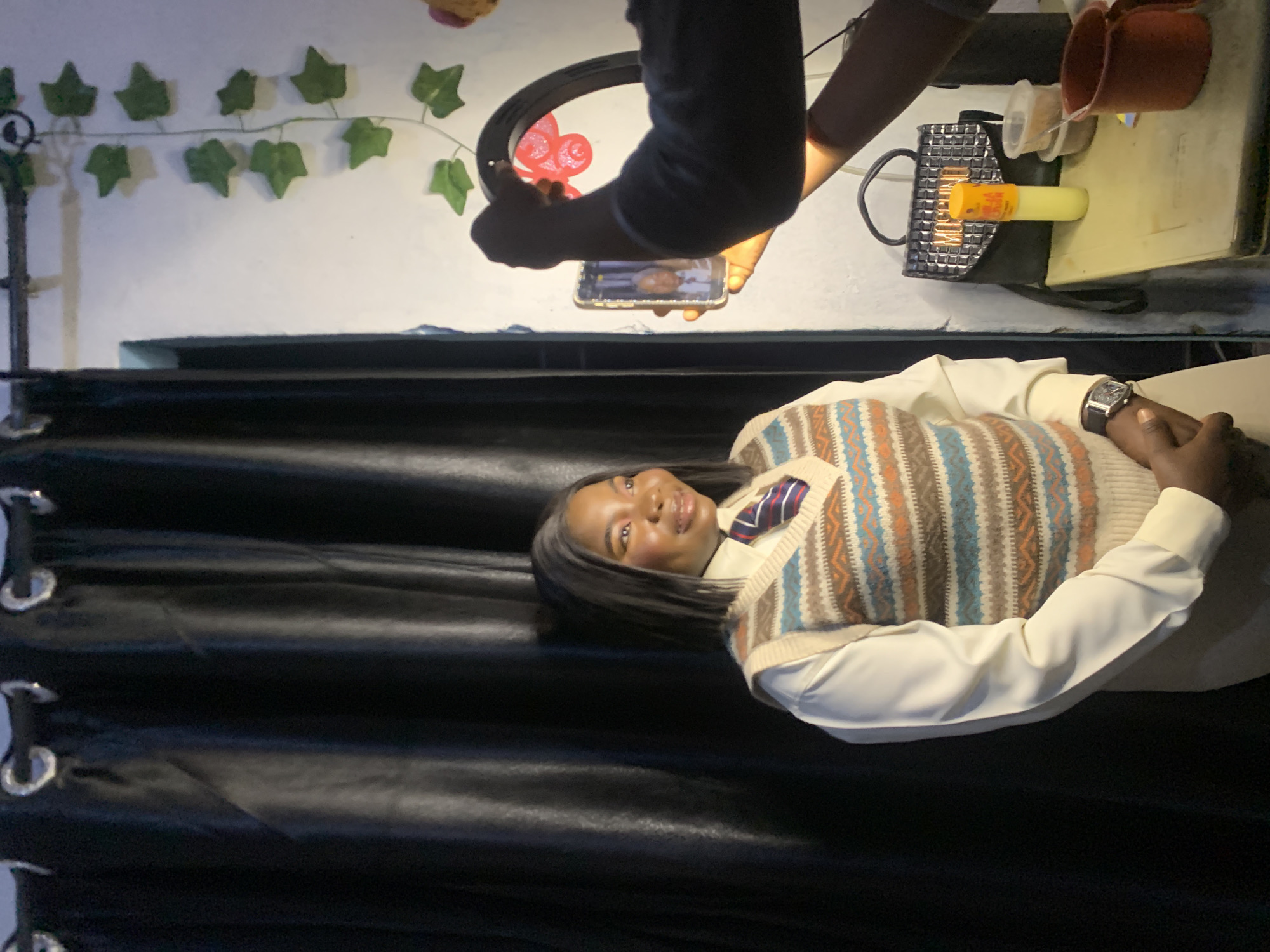 The image shows a young Black woman posing for a photograph in what appears to be an indoor setting. She is wearing a light-colored collared shirt with a patterned sweater vest over it and a striped tie. She has long, dark hair and is smiling at the camera. The background features a black curtain and a white wall adorned with artificial green ivy. In the upper portion of the image, a person's arms are visible holding a smartphone, seemingly taking the picture. A ring light is also present, positioned above the phone. To the right of the phone, there are a few objects on a light-colored surface, including a small red cup, a clear plastic container, and a small, textured bag with a yellow cylindrical object. The lighting suggests it is daytime, but the indoor setting makes it difficult to ascertain specific weather conditions. The overall mood appears cheerful and focused on capturing a portrait. The location is identified as Gwafan, Nigeria.