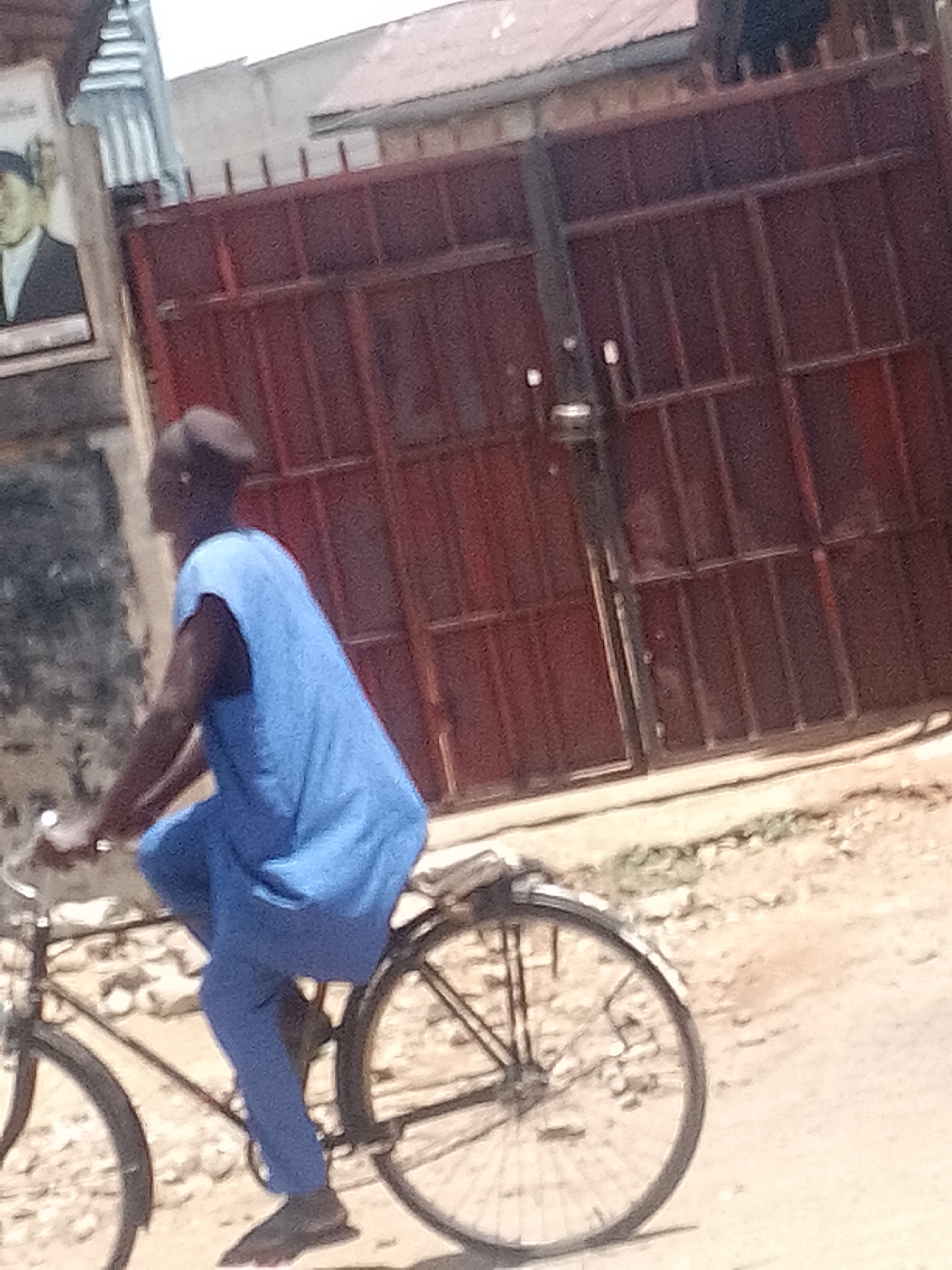 The image depicts a person riding a bicycle outdoors on a sunny day in Pantame, Nigeria. The main subject, seen from the side, is dressed in a light blue, loose-fitting sleeveless top and matching pants, with dark footwear. Their head appears to have a close-cropped hairstyle or cap. They are actively riding a dark-colored bicycle, with the frame and rear wheel clearly visible as they move from left to right across the frame.
In the background, a prominent feature is a large, reddish-brown metal gate with a grid-like structure, likely marking the entrance to a property. Behind the gate, parts of a light-colored building with a roof are visible. To the far left of the frame, affixed to a wall, is a framed portrait of a man in a suit. The details of the portrait are indistinct, and any accompanying text is illegible. The ground beneath the bicycle and in the foreground is a light-colored, dusty surface, suggesting a dirt or gravel path. The bright illumination indicates it is daytime, likely under clear, sunny skies.