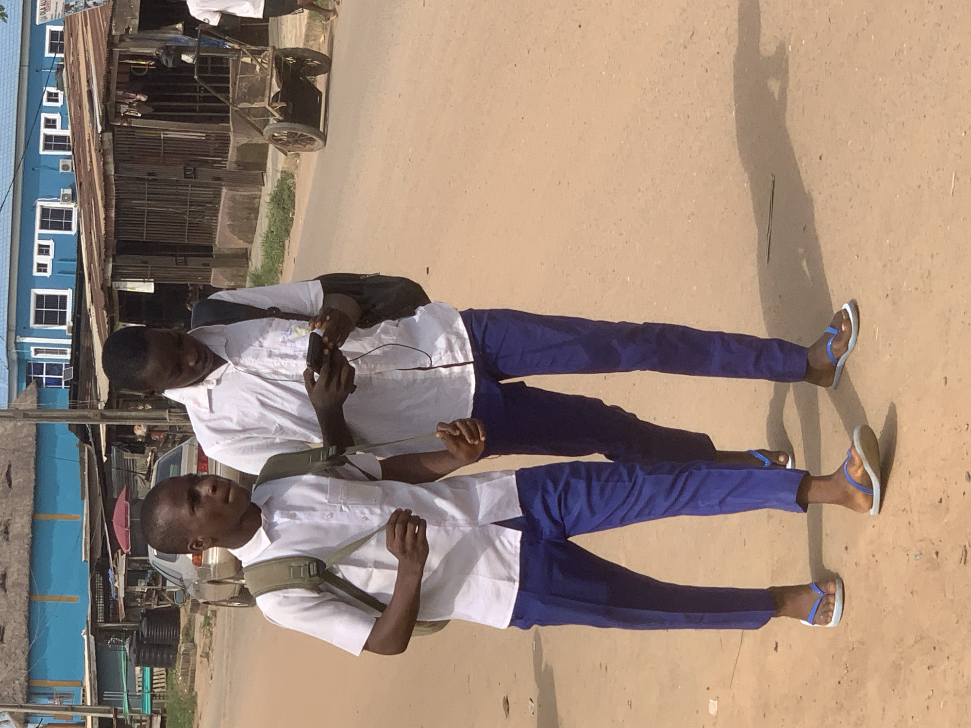 Two young men, likely students, are standing on a street in Shagamu, Nigeria. They are wearing similar white short-sleeved shirts and blue trousers with backpacks and blue/white flip-flops. One of the men is holding a mobile phone in his hand. The street appears to be paved but dusty. Behind the men, there is a mix of buildings, including a blue two-story structure with multiple windows. Various objects like a cart, a car, and water tanks are visible in the background.