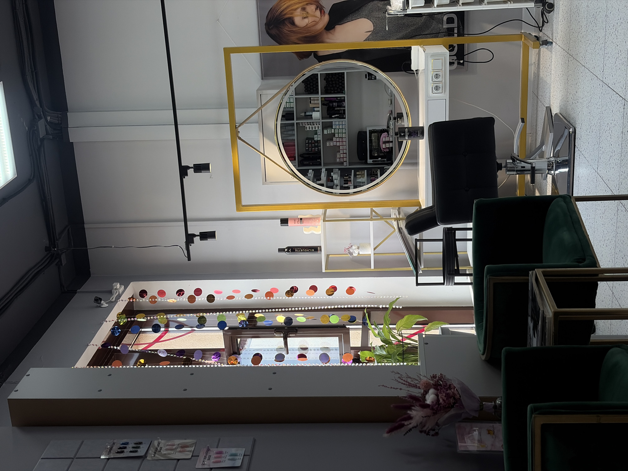 The image shows the interior of a brightly lit, modern hair salon, possibly located in Saint Petersburg, Russia. Sunlight streams in through a window, illuminating colorful beaded decorations hanging in front of it and a leafy green plant in a white pot. To the right, a large circular mirror with a golden frame is mounted on a white vanity, reflecting shelves stocked with various hair products and equipment. A black hydraulic styling chair with a chrome base is positioned in front of the vanity. In the foreground, two dark green velvet chairs with gold trim face a small side table with a magazine on it. A woman with short, reddish-brown hair is partially visible to the right of the mirror, looking down and to her left. The walls are painted white and grey, and the floor is tiled with a speckled pattern. The overall impression is one of a clean, well-organized, and stylish salon space.