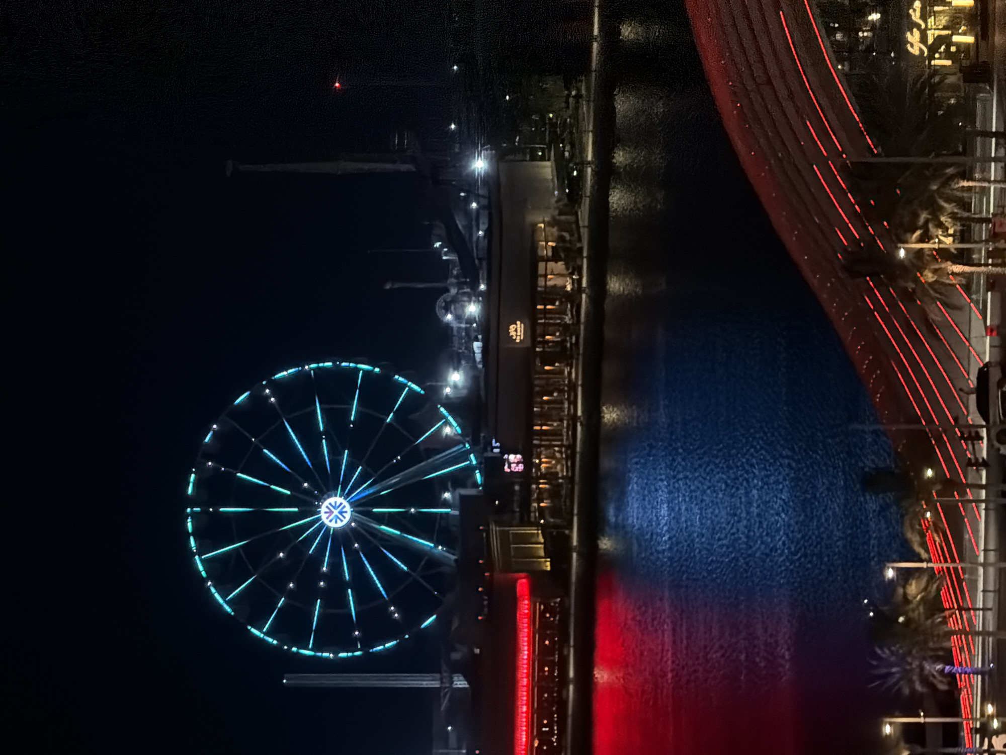 The image depicts a nighttime scene of a Ferris wheel illuminated with blue and green lights, situated in the upper left portion of the frame. Below the Ferris wheel, a body of water reflects red and blue lights from structures on the shore. Several buildings with illuminated windows are visible along the waterfront, some featuring red lighting. In the foreground, palm trees and streetlights line a waterfront promenade, and a distinctive red, arched structure with diagonal red lighting extends from the lower right corner across the frame. The background is dark, suggesting a clear night sky, with some industrial structures and tall poles visible to the right of the Ferris wheel.