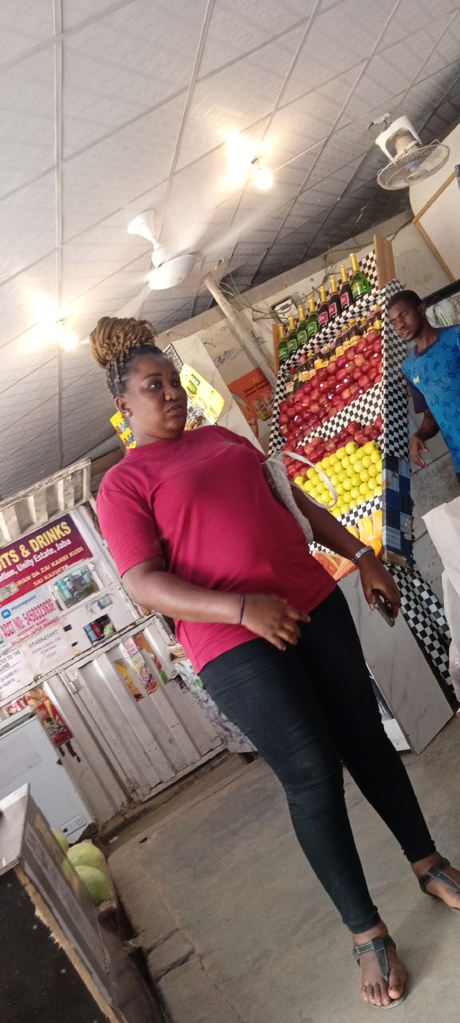 The image depicts an indoor market scene, possibly a fruit and drinks stall, in Aviation Quarters, Nigeria. A woman with her hair styled in a bun is the primary subject, seen from the chest down, wearing a red t-shirt and black pants. She is standing to the left of the frame. To her right, a man wearing a blue t-shirt is visible. The background features shelves stocked with rows of red apples and yellow citrus fruits, with green and red bottles of beverages displayed above. A checkered pattern adorns the display shelves. A white ceiling with visible light fixtures and ceiling fans is overhead. The ground is a concrete floor. Signs are visible on the stall, indicating "FRUITS & DRINKS" and other text that is partially obscured. A white cooler is also part of the stall's structure. The overall impression is a bustling, open-air market environment.