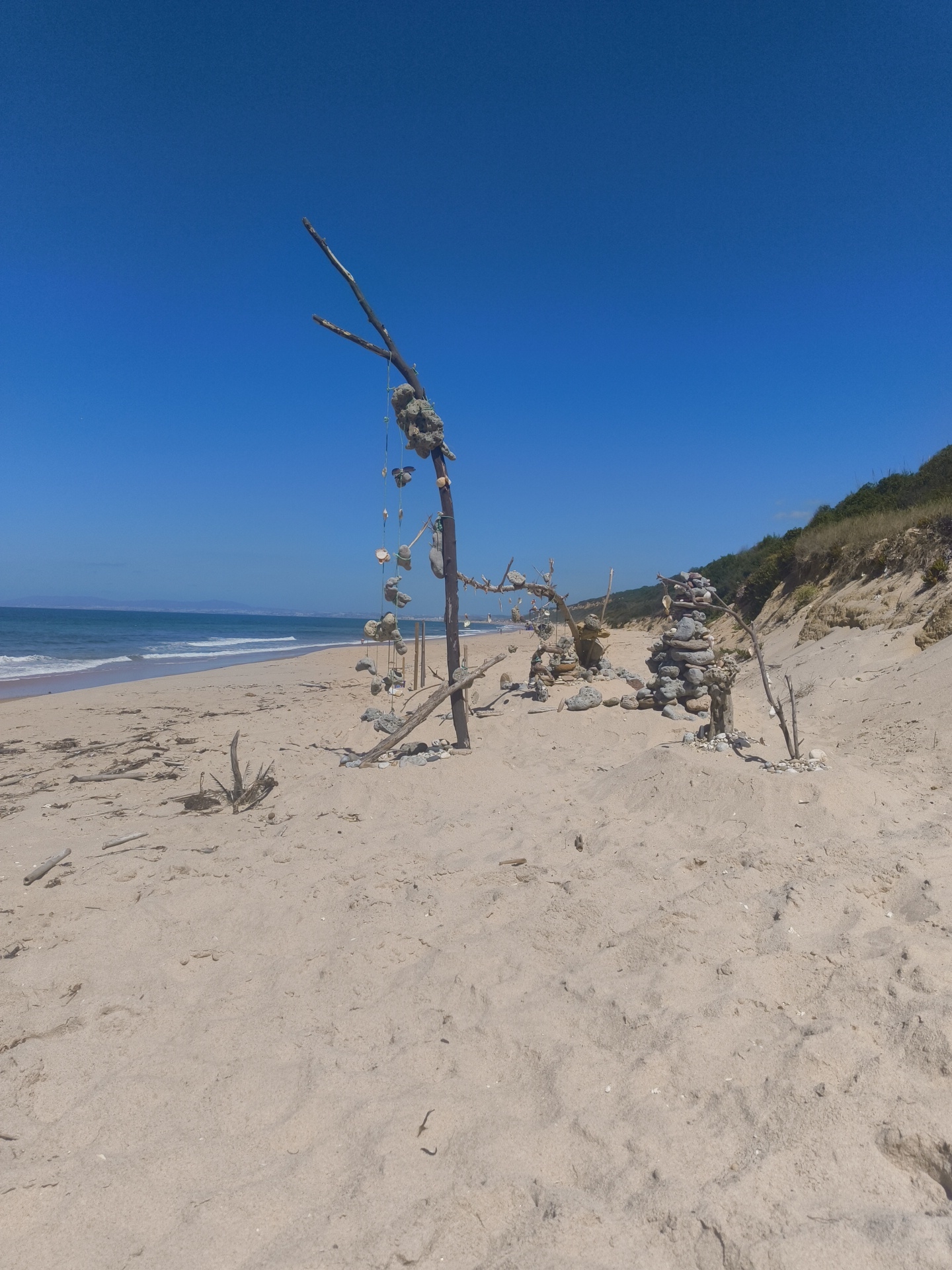The image depicts a sandy beach with a clear blue sky. In the foreground, a wooden structure with stones and shells hanging from it stands on the sand. To the right of this structure, there is a tall stack of stones. Further to the right, a grassy hill slopes down towards the beach. In the background, the ocean meets the horizon, with a distant coastline visible. The scene is devoid of people. The location is the Praia do Ouro beach, near Sesimbra, Portugal.