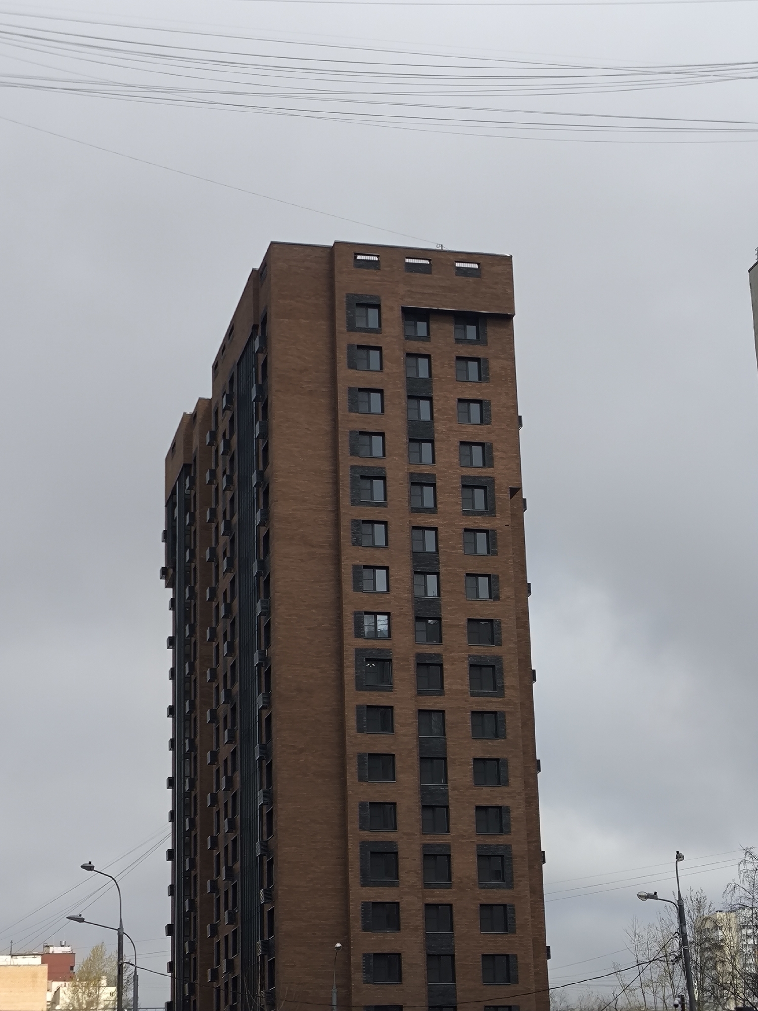 The image shows a tall, brown brick building with many windows, likely an apartment complex. The sky is overcast. Electrical wires are visible across the top of the frame. Streetlights are present in the foreground, along with other buildings in the distance, suggesting an urban environment. Based on the metadata information, the location is Moscow, Russia.