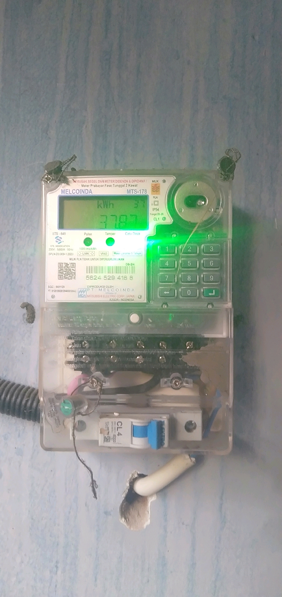 A pre-paid electricity meter from MELCOINDA, model MTS-178, is mounted on a blue-painted wall. The meter displays "kwh" and the number "3787" in green digital font. Below the display, there are indicators for "Pulse," "Tamper," and "Catu Daya." A keypad with numbers 0-9 and directional arrows is visible to the right of the display. The meter has an ID number "5624 529 418 8". The lower section of the meter shows electrical connections, a small circuit breaker labeled "CL4," and a QR code. The meter is attached to the wall with visible wires and some electrical tape. A corrugated pipe enters the meter from the left side. The surrounding environment appears to be an indoor setting with a textured blue wall. The scene is located in Plemahan, Indonesia.