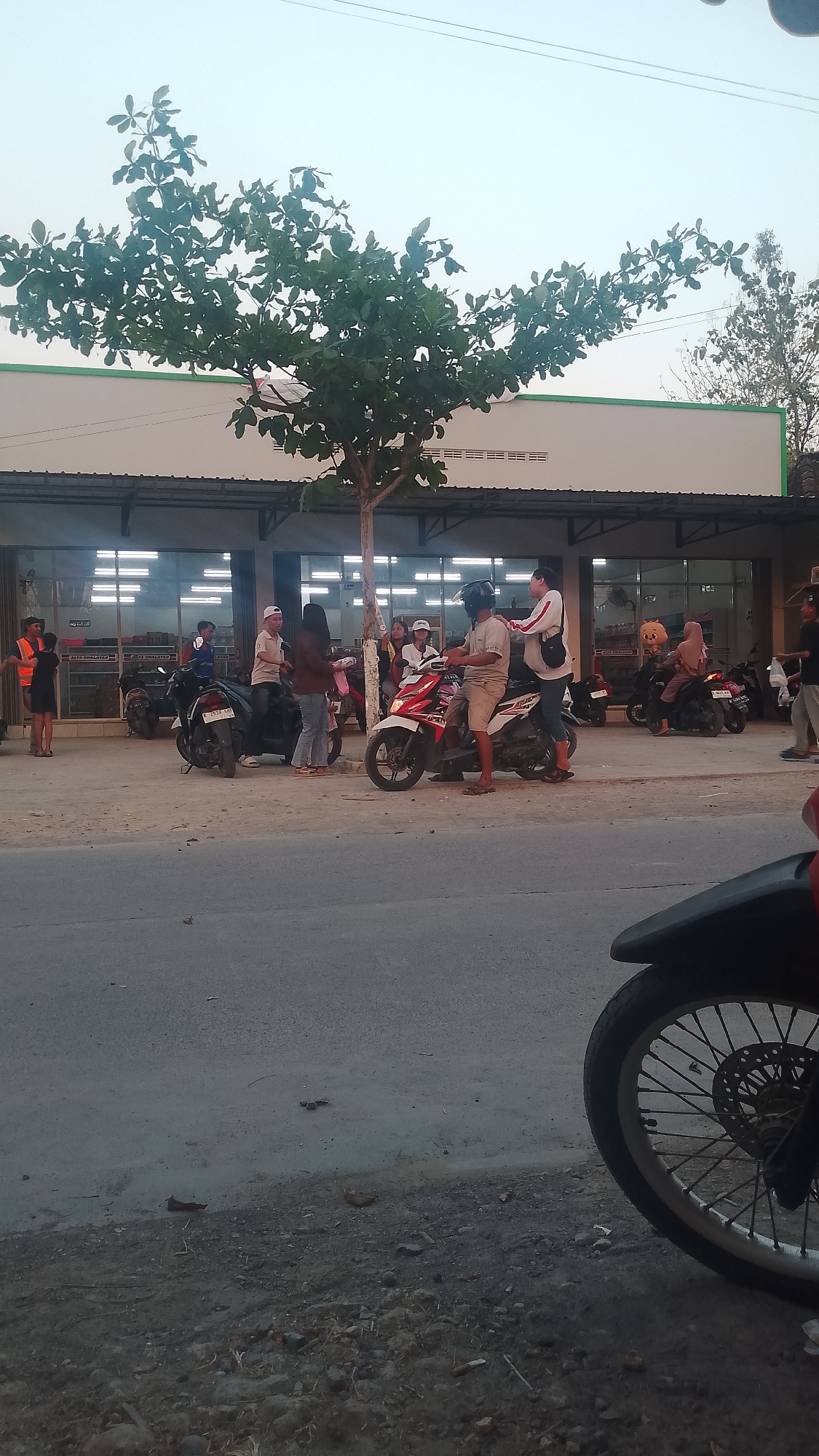 In Karanglangu, Indonesia, a street scene unfolds in front of a building with a light-colored facade and a dark awning. A tree with dense green foliage stands prominently in the foreground, casting shadows. Several people are gathered, some near motorcycles. One individual wearing an orange vest is visible on the left. A red and white motorcycle is parked in the center with people standing around it. Another group of individuals is positioned near motorcycles on the right side of the scene. The ground is unpaved, with gravel and debris. A portion of a motorcycle wheel and part of its fender are visible in the lower right foreground, suggesting a low-angle perspective or another vehicle present. The sky is a pale, hazy blue.