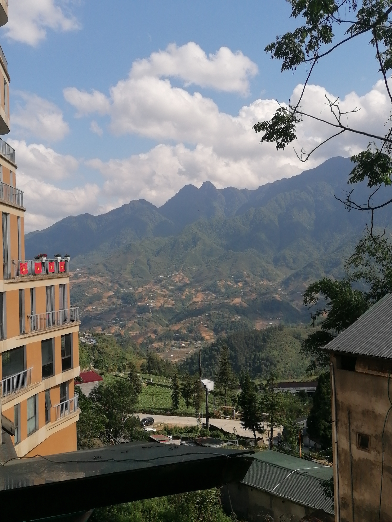 The image shows a mountainous landscape in Sa Pa Ward, Vietnam, under a partly cloudy sky. A multi-story building, possibly a hotel or apartment complex, is situated on the left, adorned with Vietnamese flags on its balconies. Terraced fields and settlements are visible in the valley between the mountains. Trees partially obscure the view from the right side. A rooftop of a building is visible in the foreground. A road with a vehicle on it and some buildings, power lines and utility poles can be seen in the middle ground.