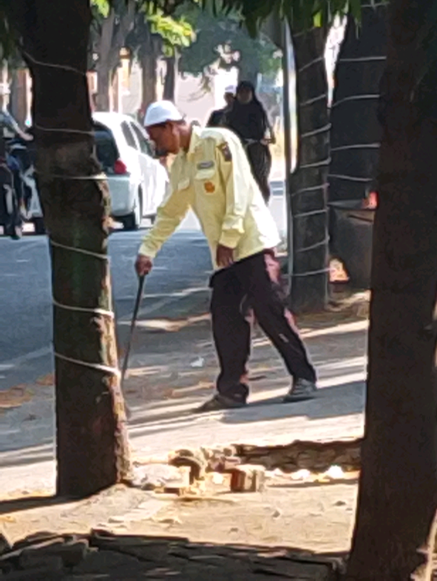 In Bendo Mungal, Indonesia, a man wearing a white cap and a yellow shirt with dark pants walks with a cane. He is positioned on a street, with a white car visible in the background. The scene is framed by trees, one of which is wrapped with white rope. The ground is a mix of pavement and dirt with scattered debris. Another person, wearing dark clothing, is partially visible in the background.