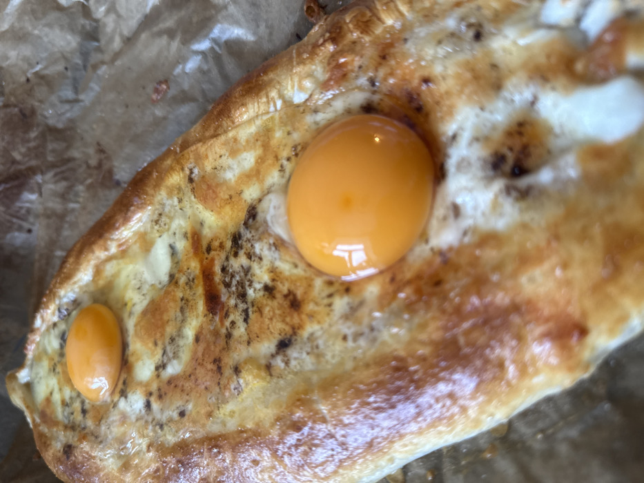 The image displays a close-up, overhead view of a boat-shaped Khachapuri Adjaruli, a traditional Georgian cheese bread with egg, located in Yekaterinburg, Russia. The baked item rests on a crumpled sheet of light brown parchment paper. Its golden-brown crust, with raised, ridged edges, surrounds a central cavity filled with a melted white cheese mixture. Within this cheese, two unmixed, glossy, bright orange-yellow egg yolks are visible: one prominent in the lower-right foreground and a smaller, partially obscured one in the upper-left background. The cheese surface exhibits light browning and dark, granular specks, consistent with herbs or seasoning. The parchment paper background shows creases and subtle tonal variations from contact with the food. No individuals or actions are depicted.