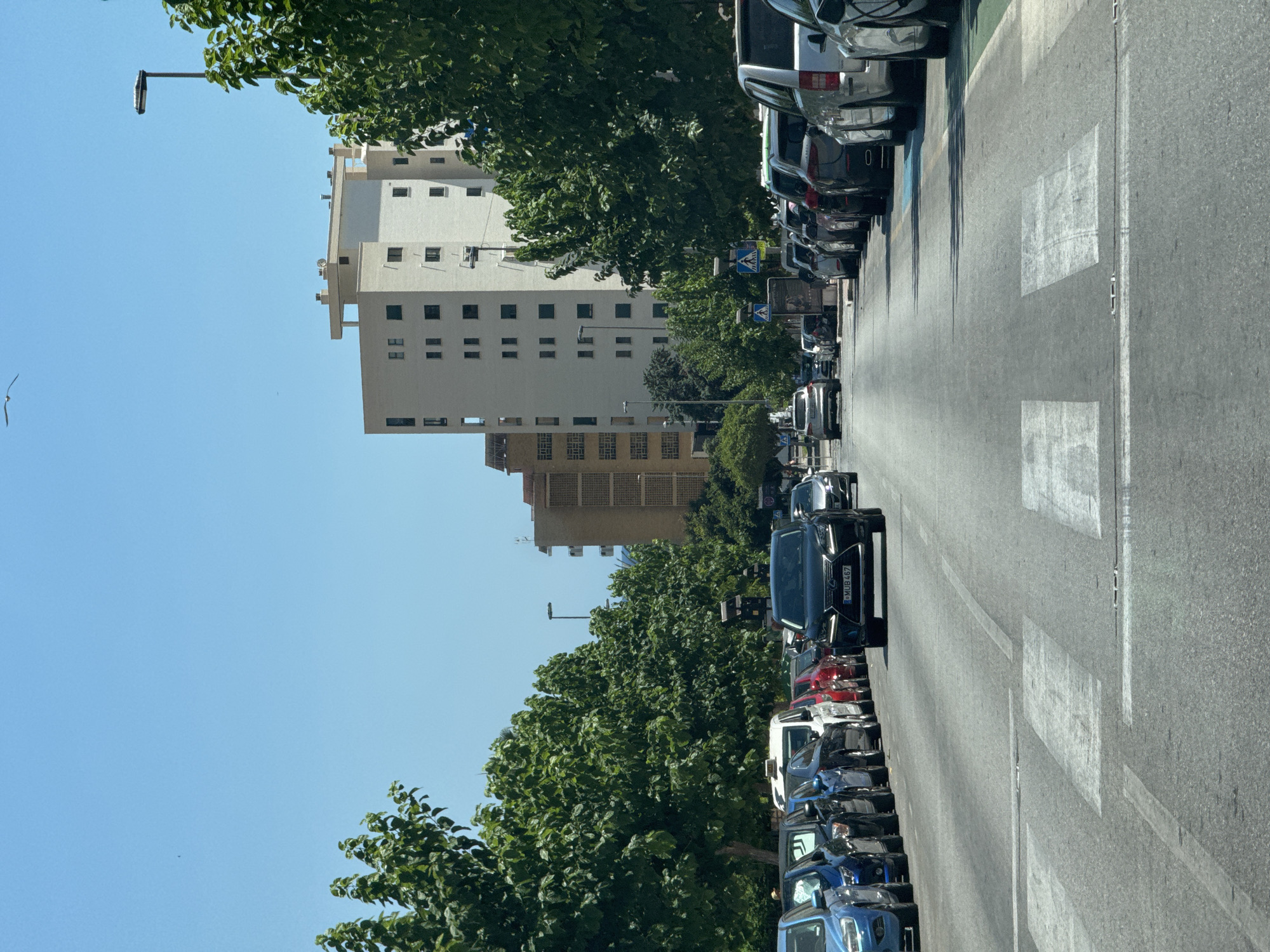 The image shows a street scene in La Vila Joiosa, Spain. A black Lexus SUV is in the foreground, stopped at a pedestrian crossing. The car has a license plate that reads "MUB 467". Behind the Lexus, a line of cars is parked on the right side of the street, and more cars are driving in the same direction. On the left side of the street, several cars are also parked. Tall, leafy green trees line both sides of the street, casting shadows on the road. In the background, several multi-story apartment buildings are visible. The sky is clear and blue. A lamppost is visible on the right side of the image. The scene appears to be a typical urban environment on a sunny day.