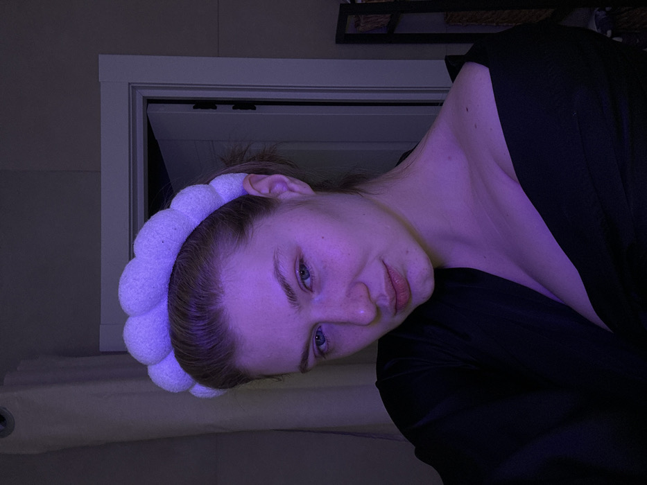 A woman is positioned at an angle, gazing towards the camera. She sports a white, fluffy headband that stands out against her hair. A dark top covers her upper body. The lighting casts a purplish hue over the scene, highlighting her fair complexion and blue eyes. A partially visible white window frame is behind her.